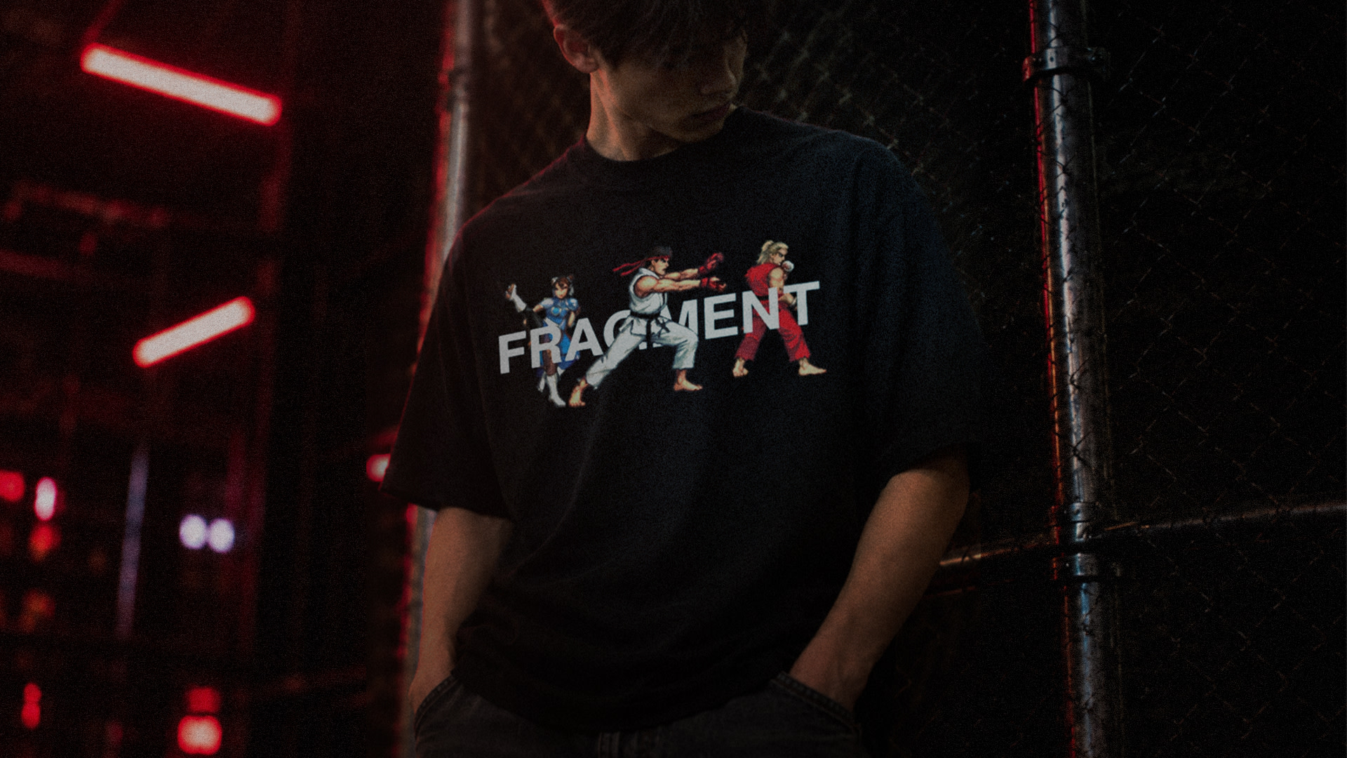 Person in a dimly lit area wearing a black T-shirt with "FRAGMENT" and characters from a fighting video game.