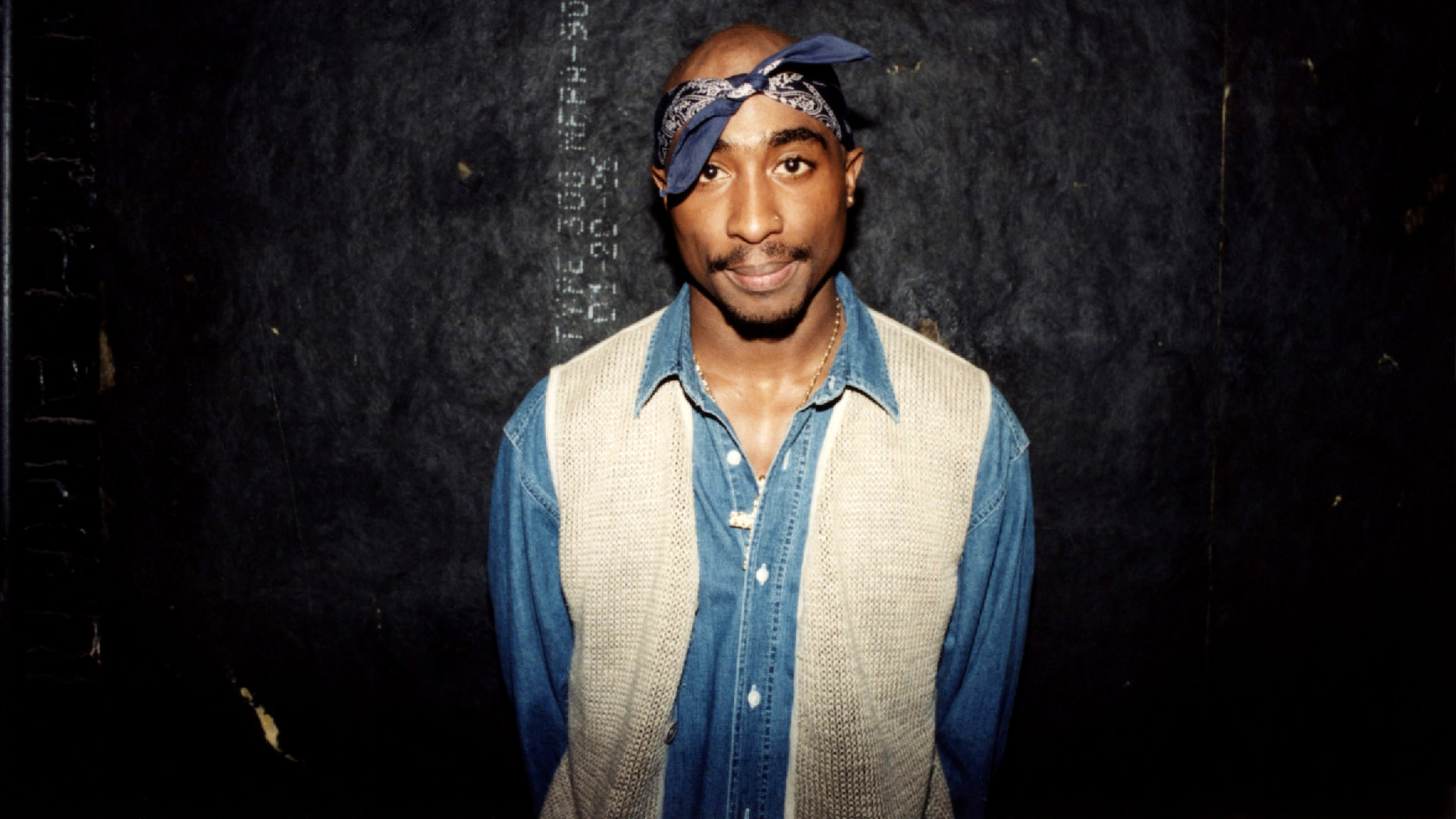 Tupac Shakur wearing a denim shirt and beige vest, with a blue bandana on his head, standing against a dark background.
