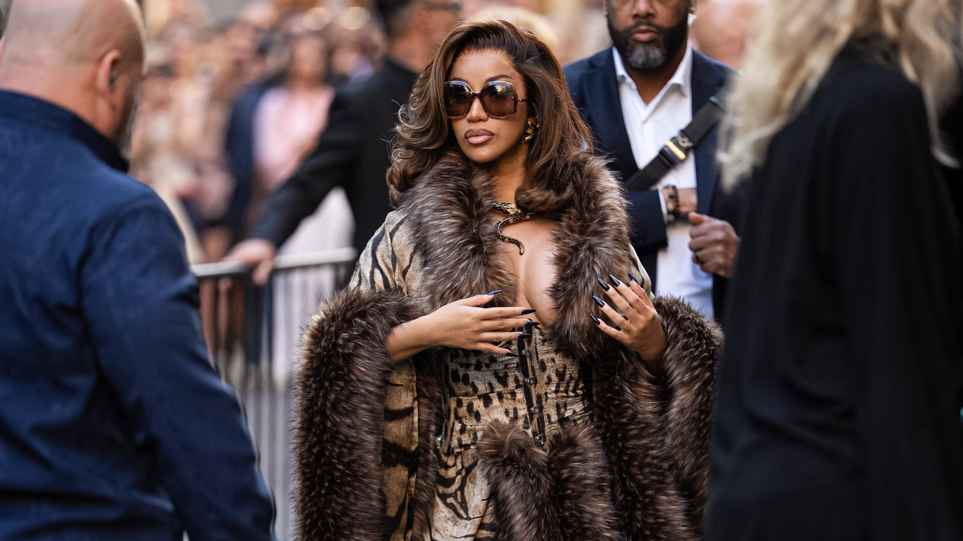 Cardi B in a stylish fur coat and sunglasses walks through a crowd. She has long hair and is surrounded by people in casual and formal attire.