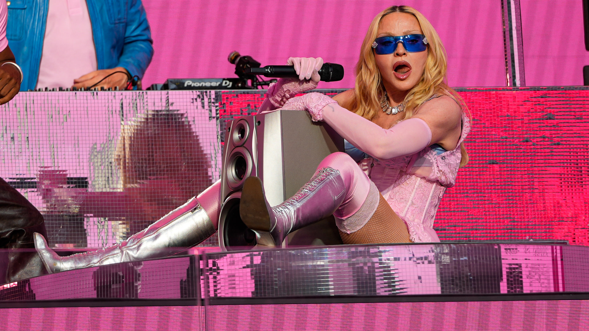Madonna in a pink corset, silver boots, and blue sunglasses sings into a microphone on a pink stage.