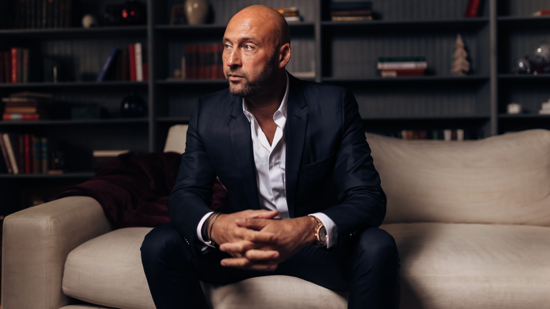 Derek Jeter in a dark suit sits on a beige couch, hands clasped, with a thoughtful expression. Bookshelves are in the background.