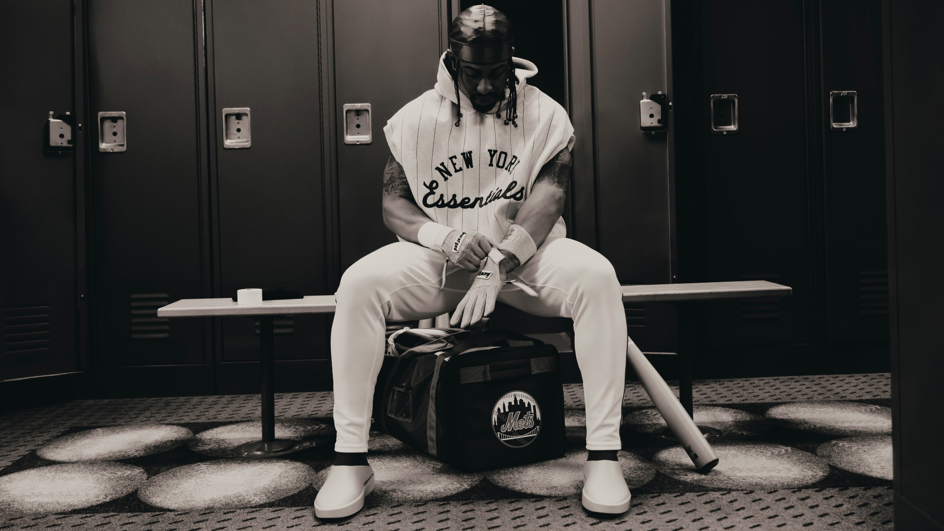 A person in a locker room sits on a bench, head down, wearing a "New York Essentials" hoodie. A bag and baseball bat are nearby.