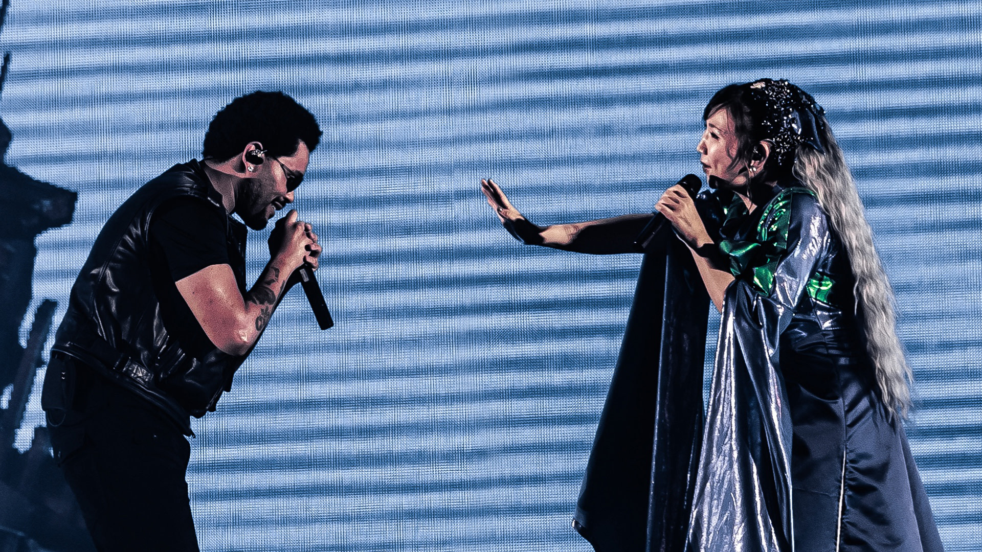 Two performers energetically singing on stage, The Weeknd, left in a black outfit, and Yoko Takahashi, right, in a metallic, flowing costume with long hair.