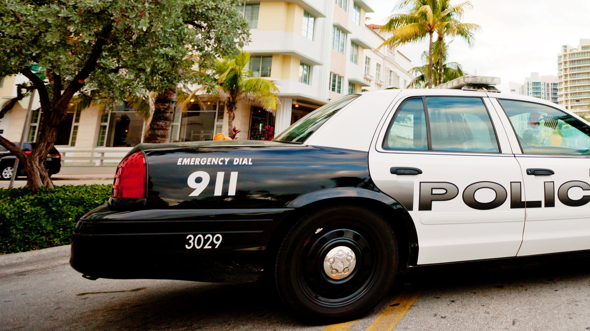 A police car parked on a street, with "911" and "POLICE" visible on the side. Palm trees and buildings are in the background.