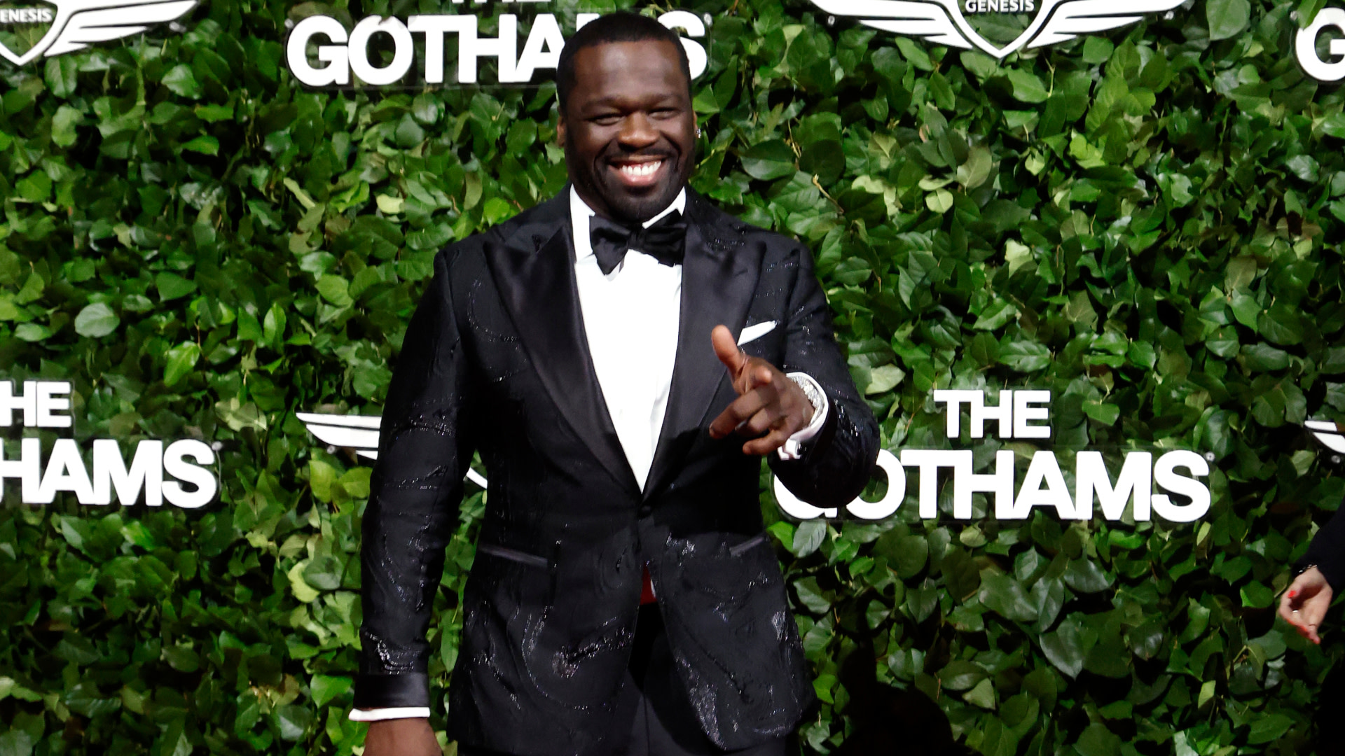 50 Cent in a black tuxedo, smiling and pointing at the Gotham Awards. Green foliage backdrop with "The Gothams" signage.