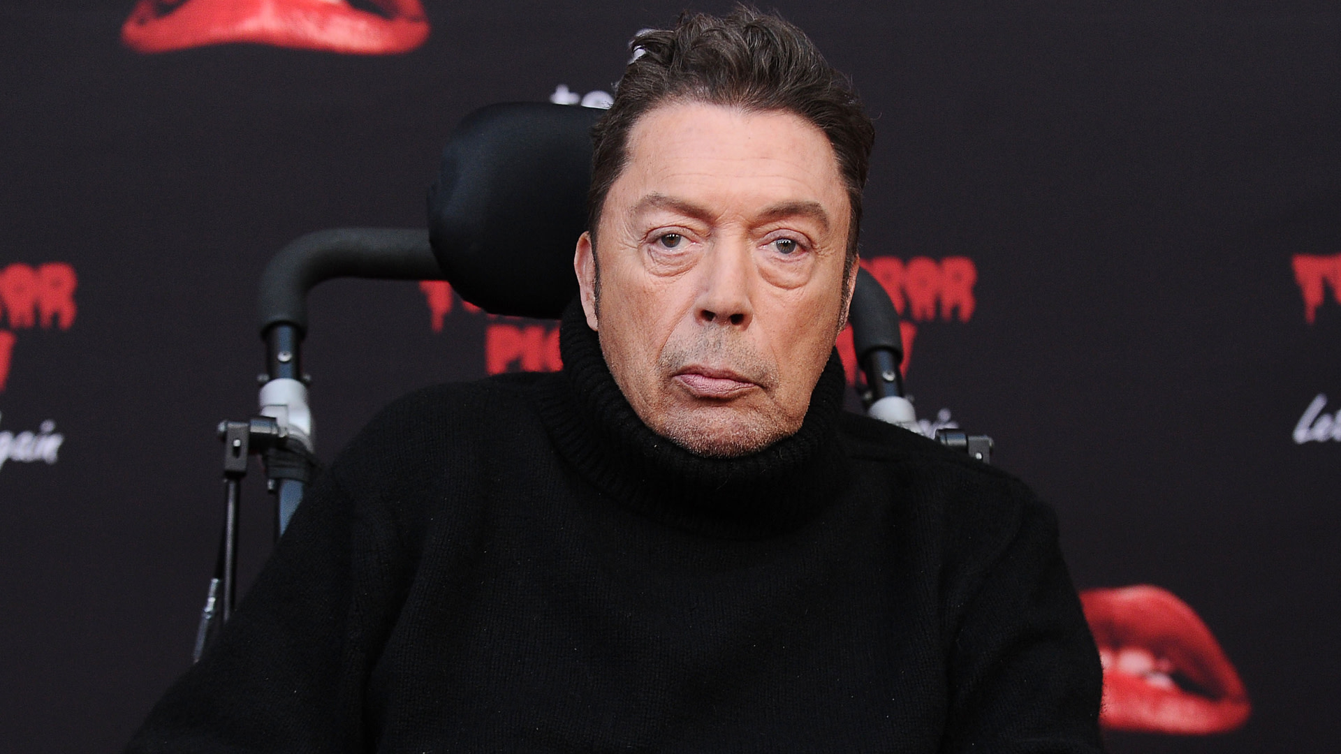 Tim Curry in a black sweater sits in a wheelchair against a backdrop with red lips and text.