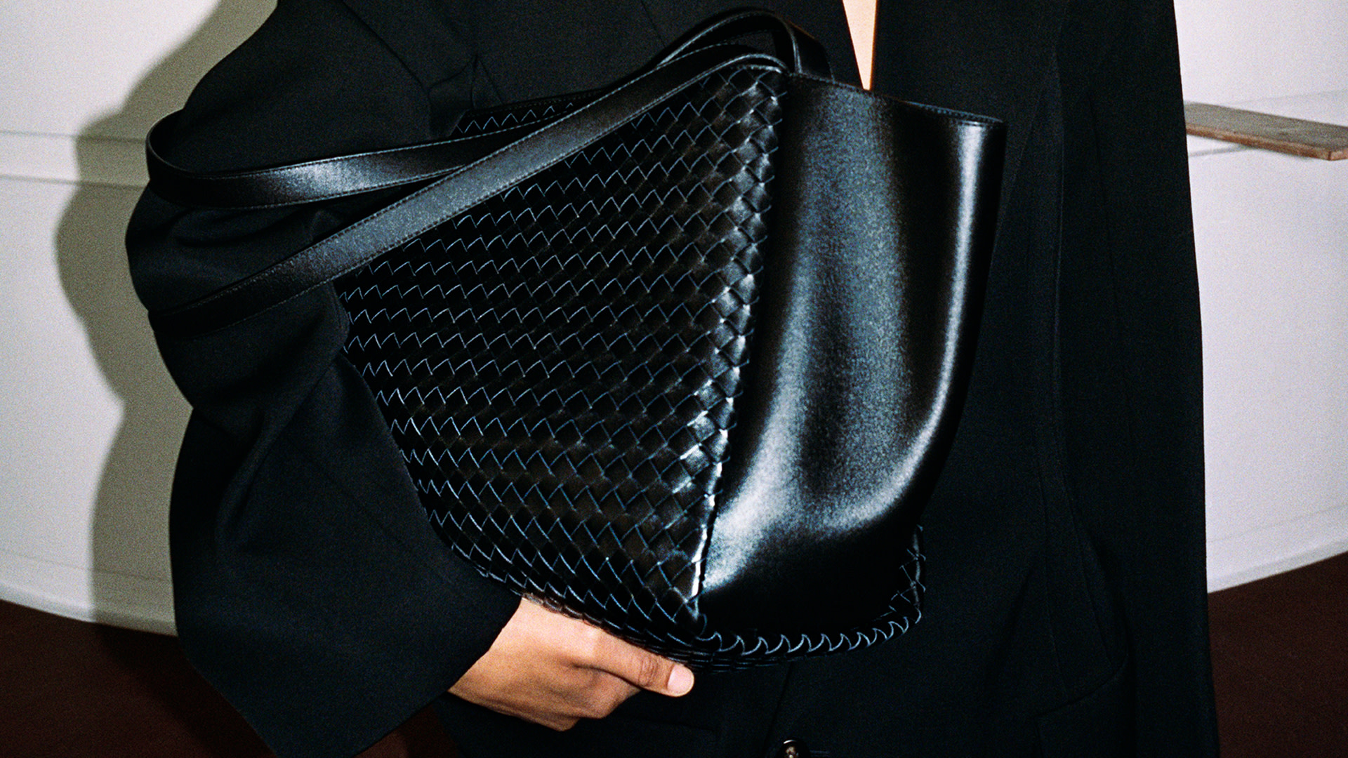A person in a black outfit holds a large black woven leather bag with a sleek design.