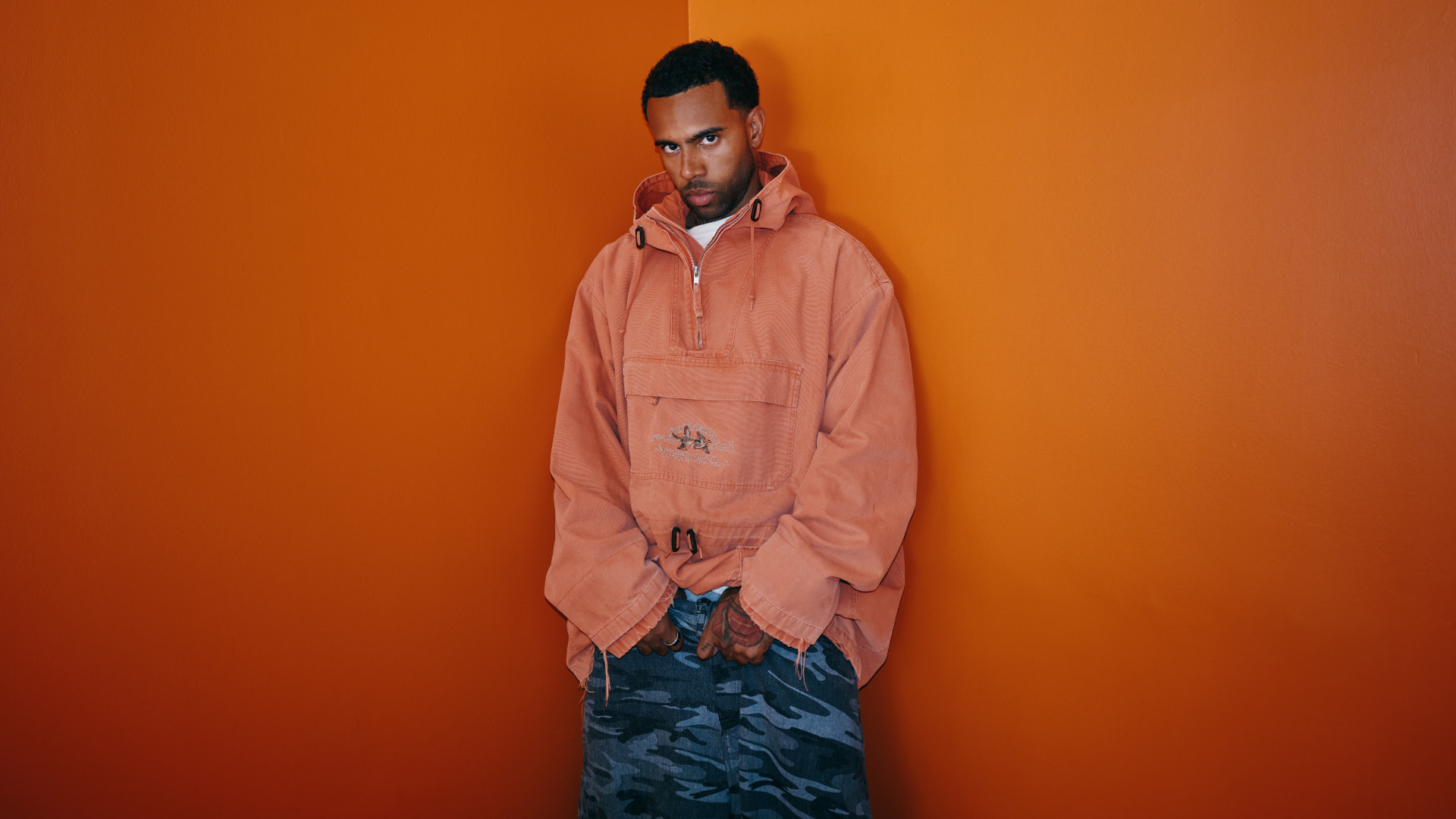 Vic Mensa in an oversized orange hoodie and camo pants stands against an orange background, looking at the camera.
