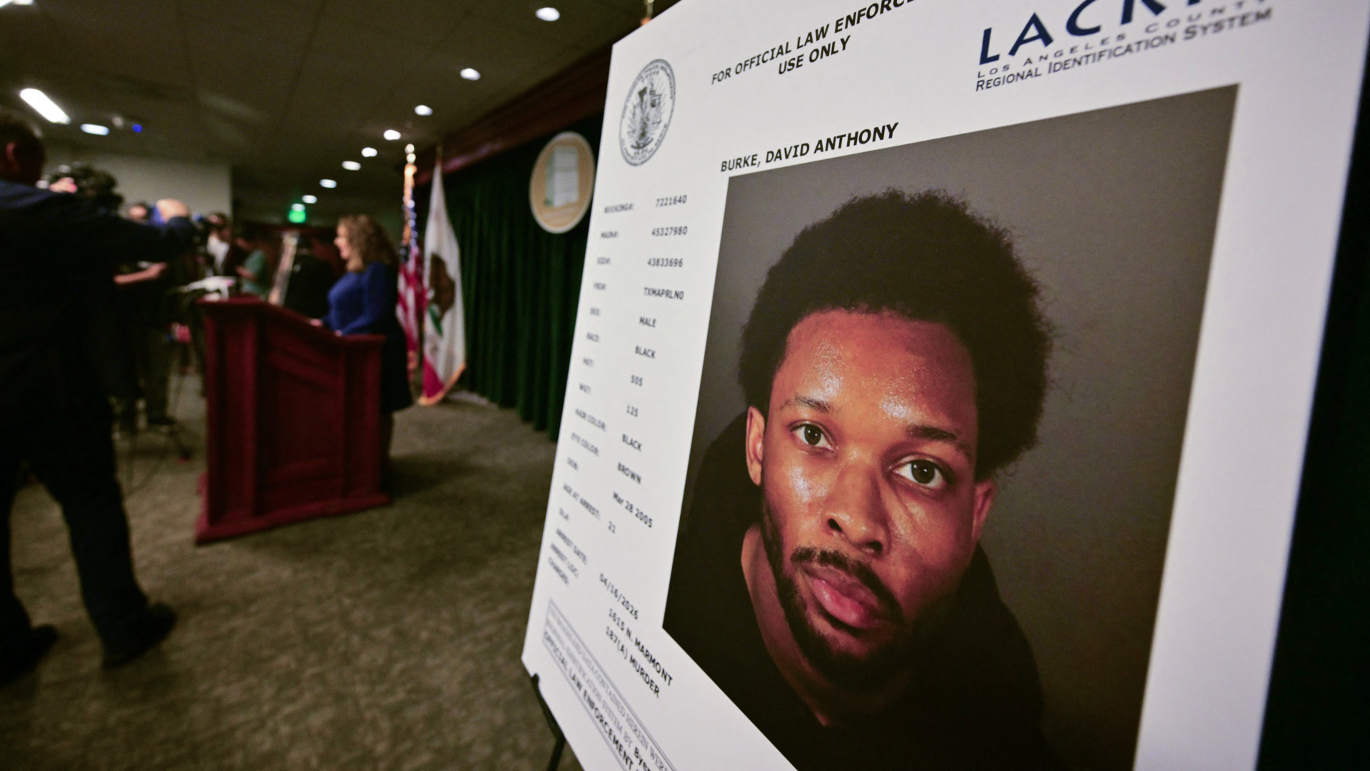 A large mugshot photo of D4vd on display at a press conference, with officials speaking at a podium in the background.
