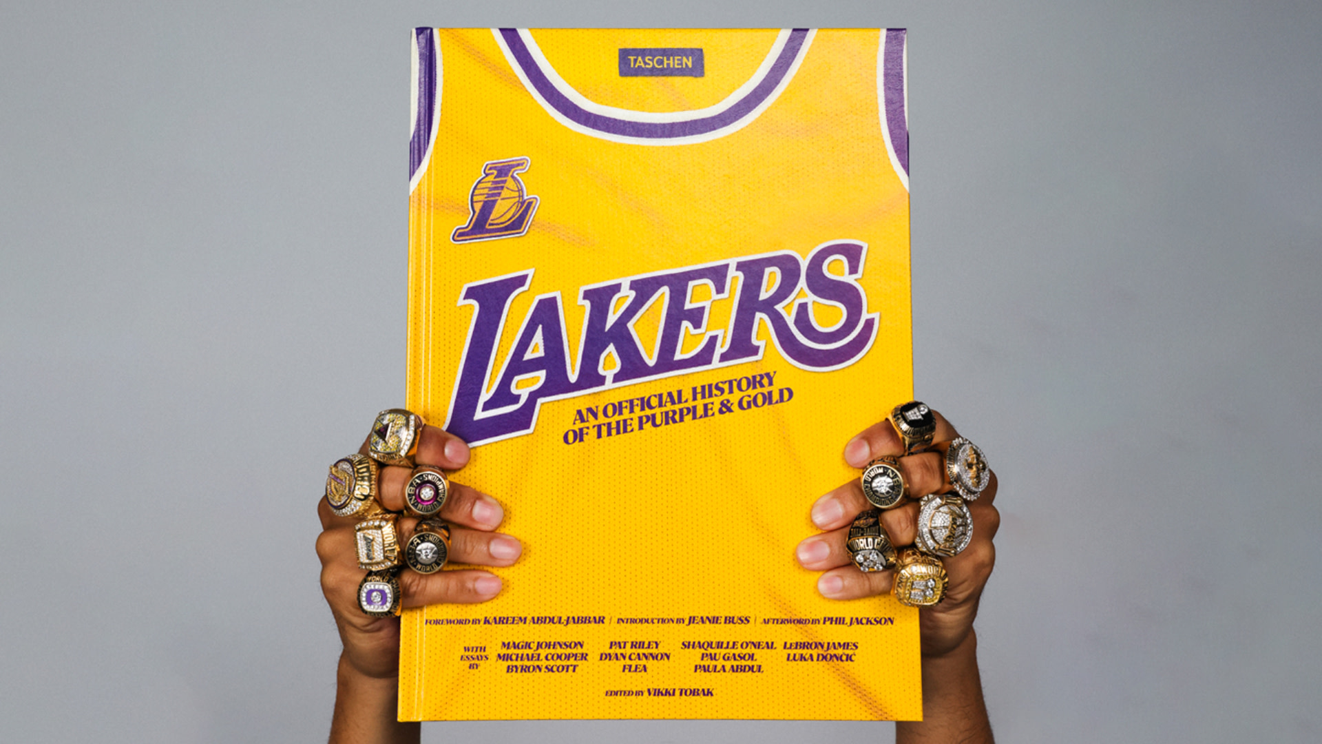 Hands with multiple championship rings hold a book titled "Lakers: An Official History of the Purple & Gold," featuring a Lakers jersey cover.