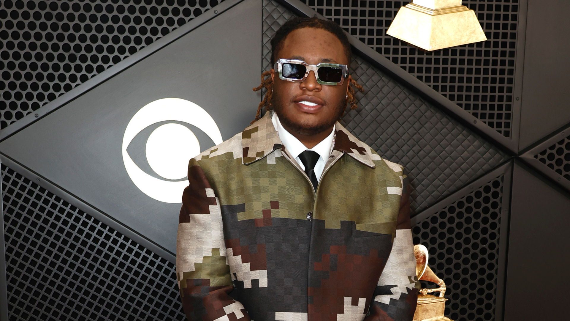 Tay Keith wearing a camouflage-patterned coat and sunglasses poses at an event with a CBS logo and Grammy trophy in the background.