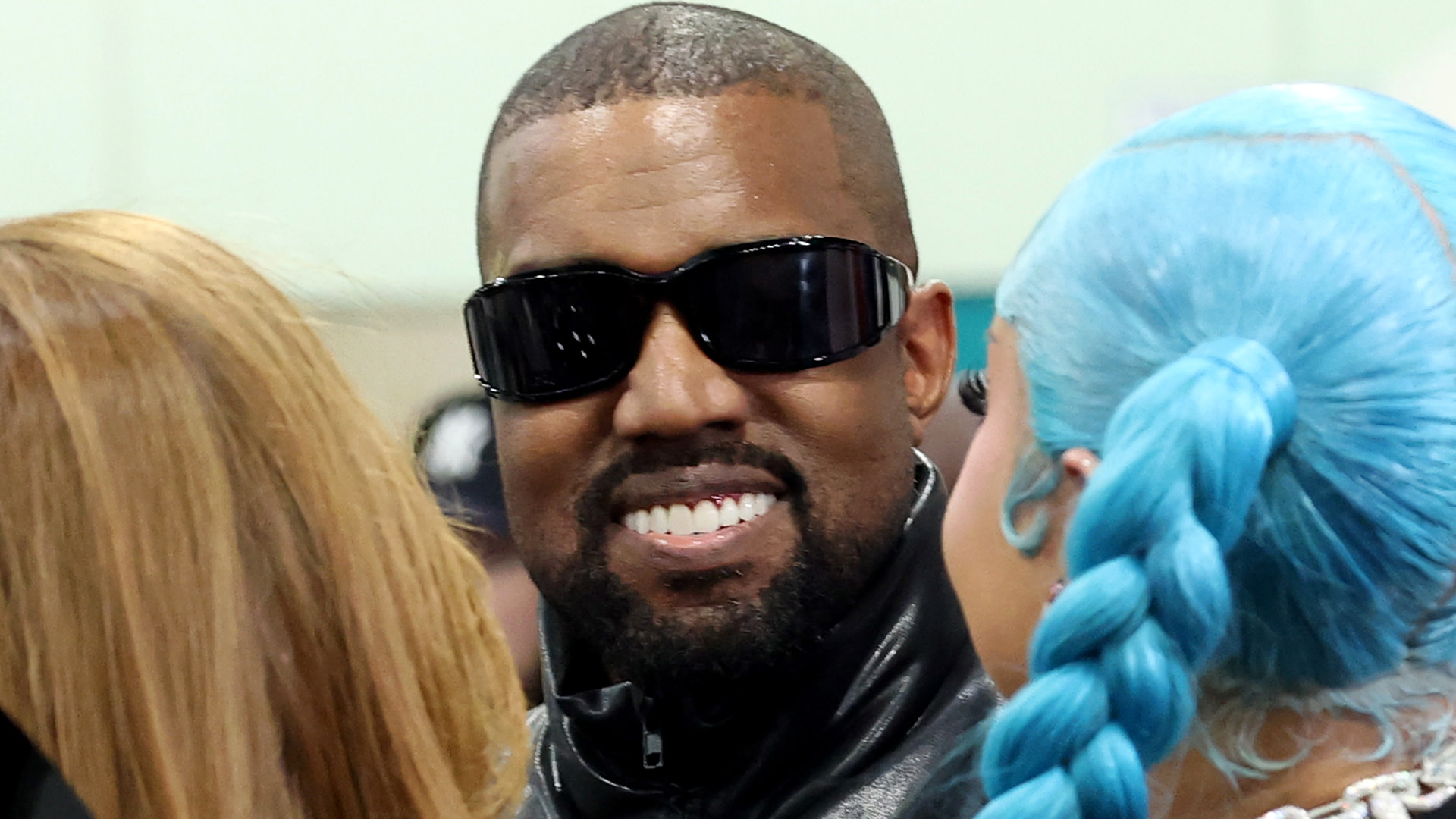 Kanye West wearing sunglasses and a black leather jacket, smiling, with a person with blue hair in the foreground.