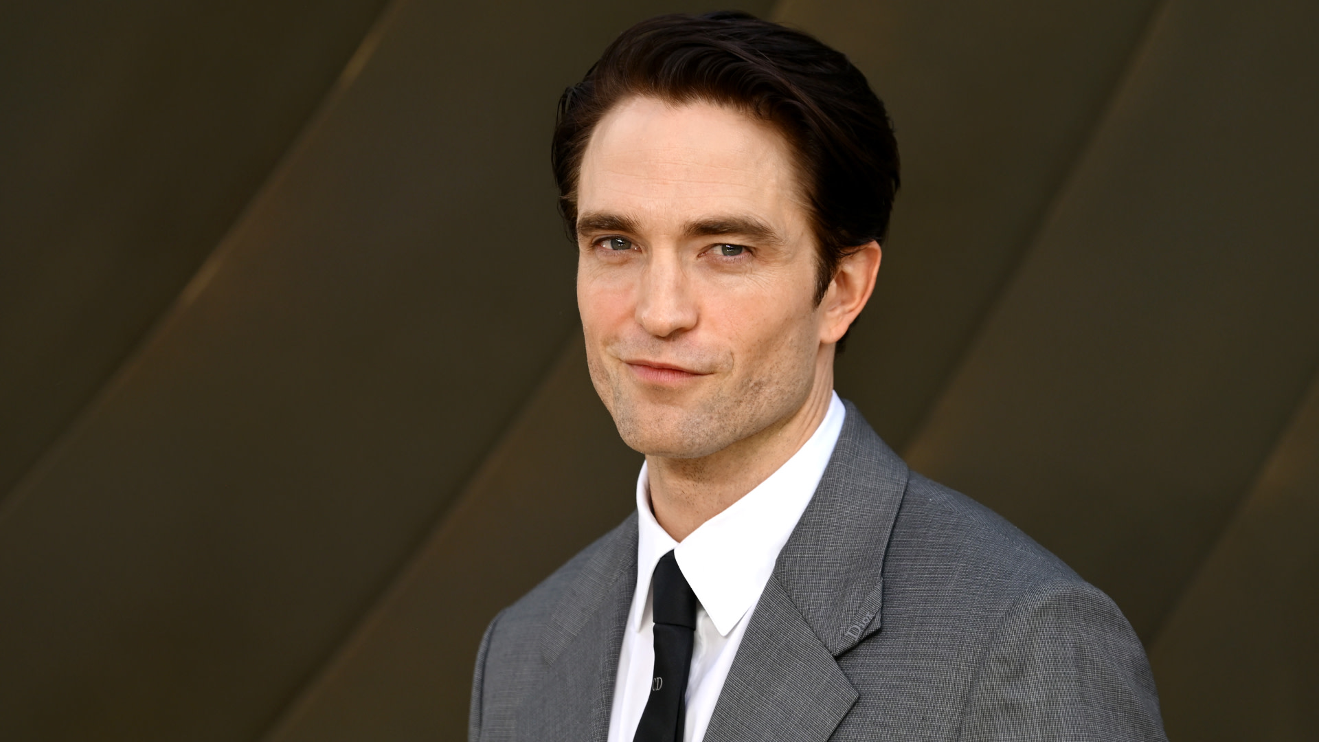 Robert Pattinson in a gray suit and black tie, standing against a dark background, with a slight smile.