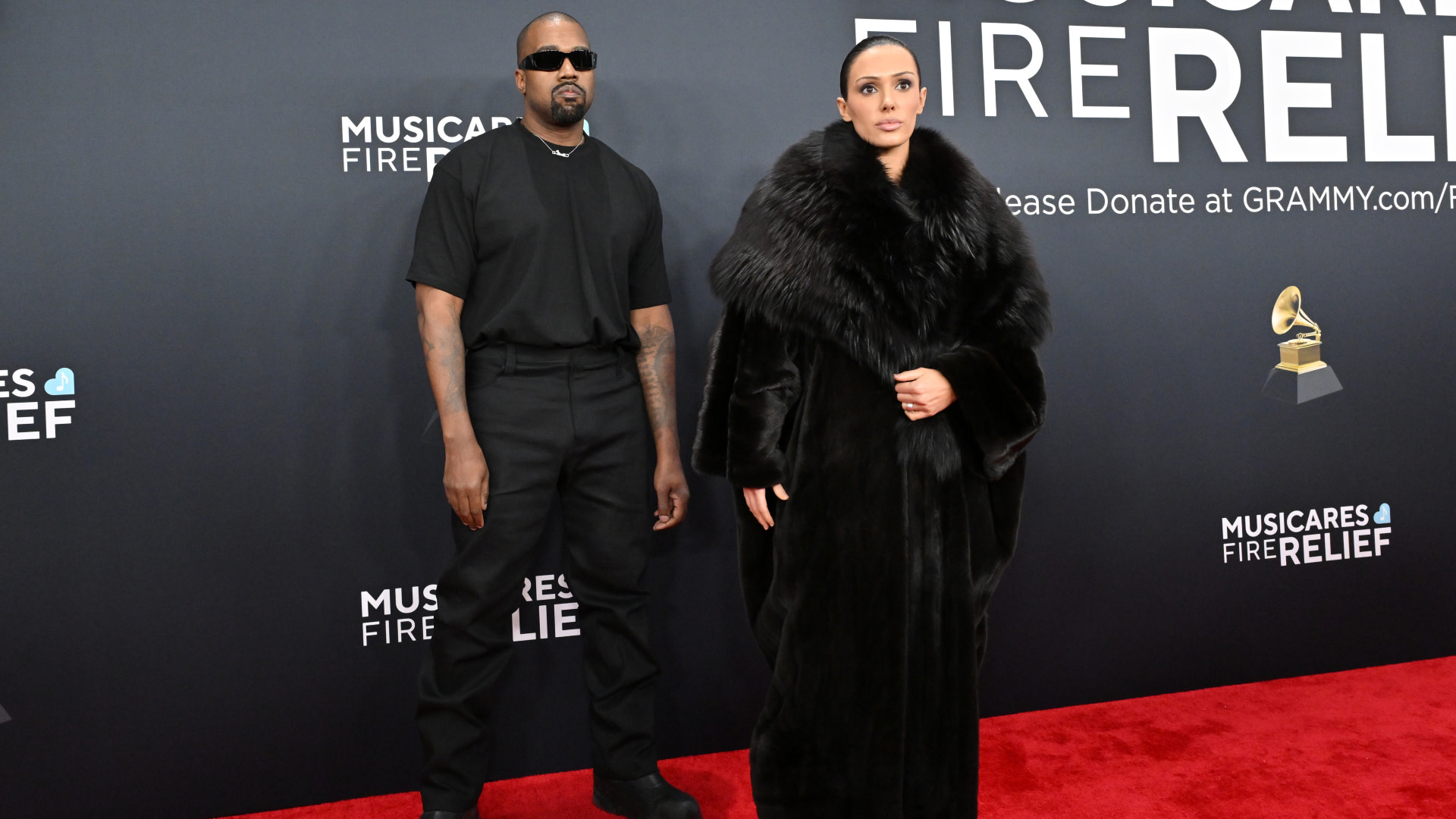 Two individuals on a red carpet at a MusiCares event. Ye wears sunglasses and black attire; Bianca Censori is in a fur coat.