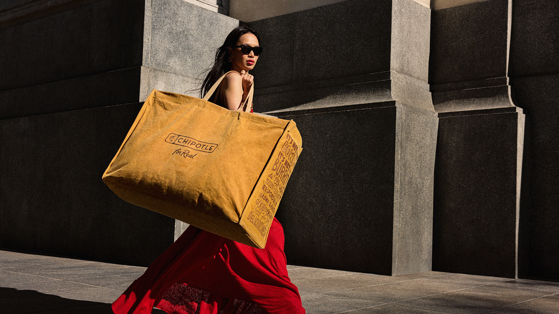 A woman in a red dress and sunglasses carries an oversized Chipotle bag on a city street.