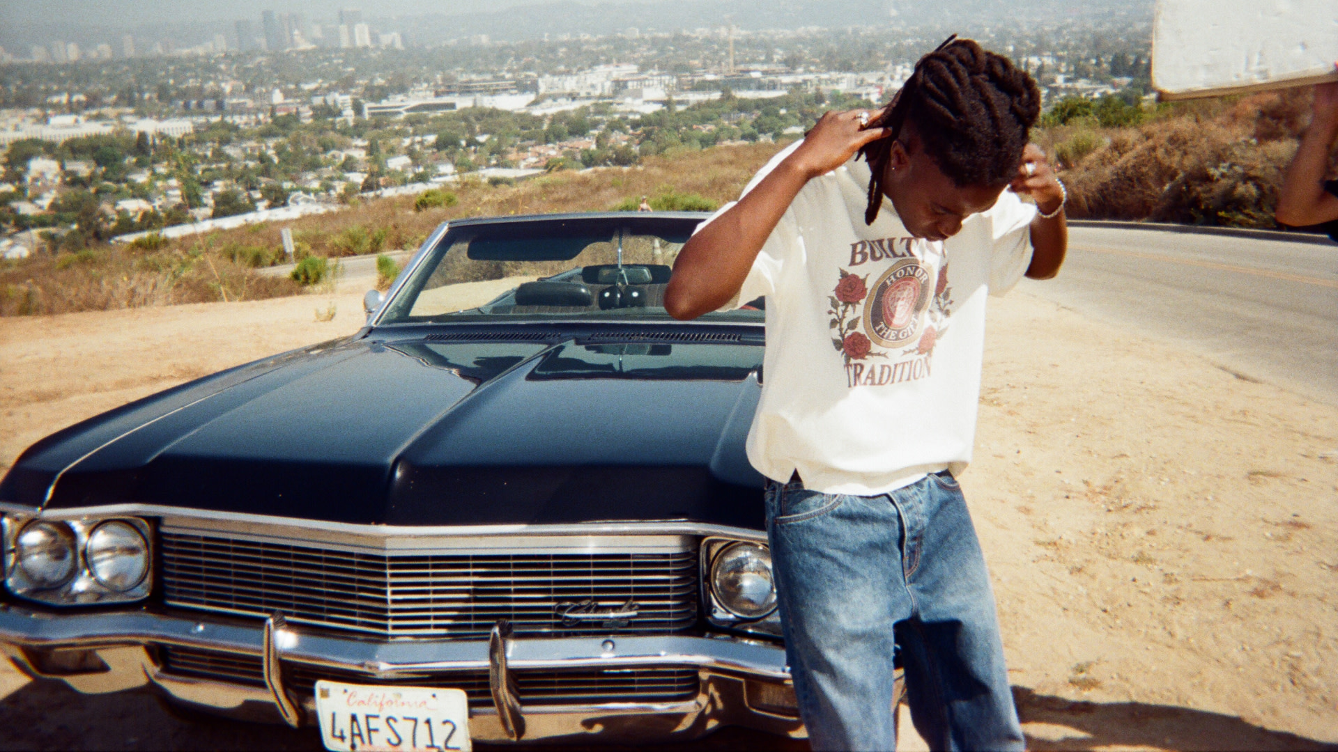 A person with dreadlocks stands in front of a classic car on a hill, adjusting their hair. Cityscape in the background.