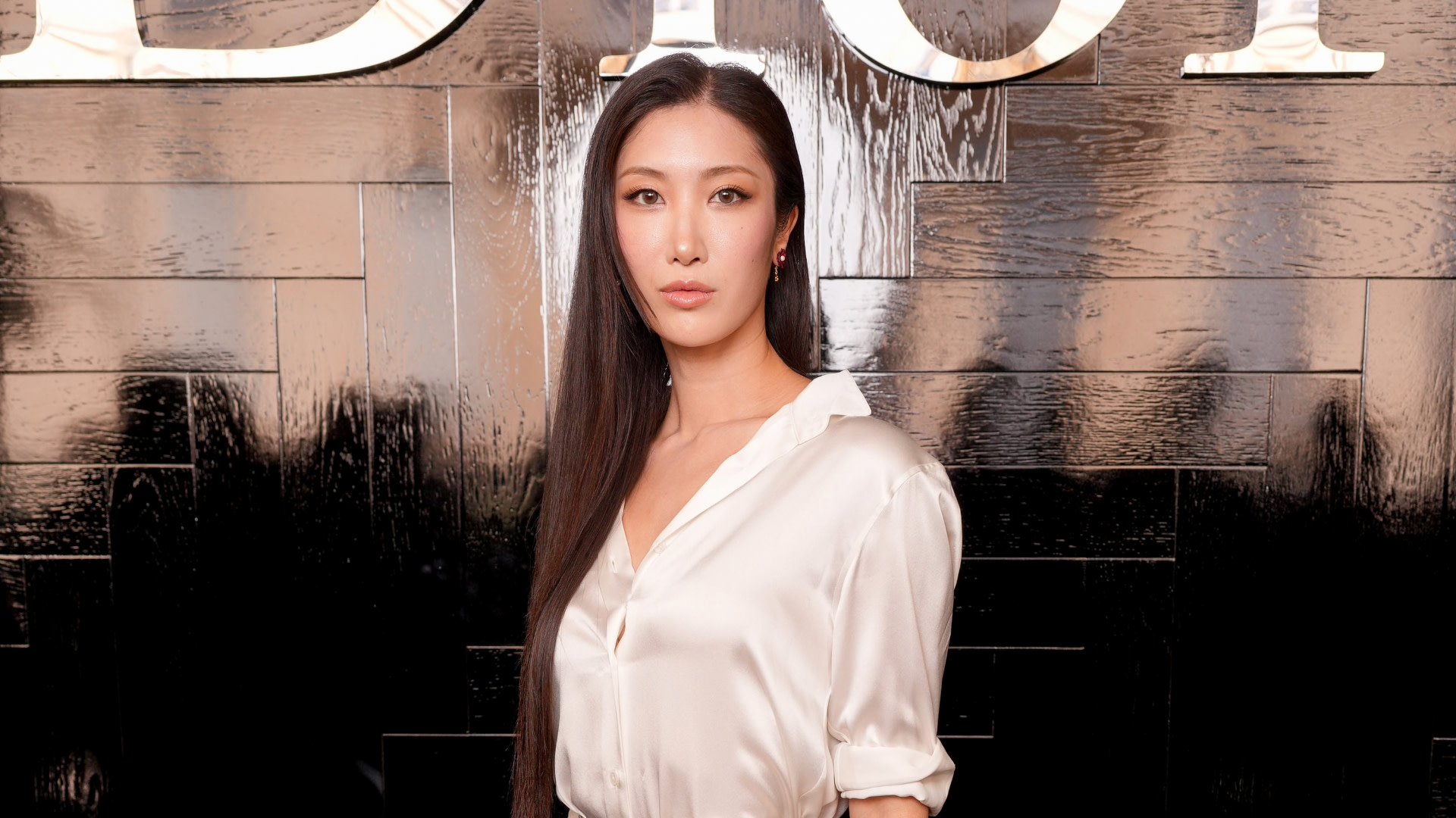 A woman with long hair in a white blouse stands against a glossy black wall with the word "Dior" partially visible above.