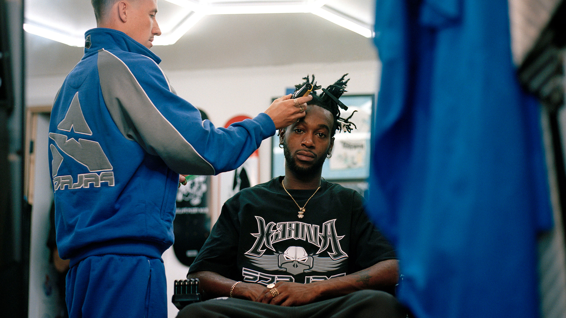 A person in a blue tracksuit styles another person's hair in a barbershop. The seated person wears a graphic T-shirt.