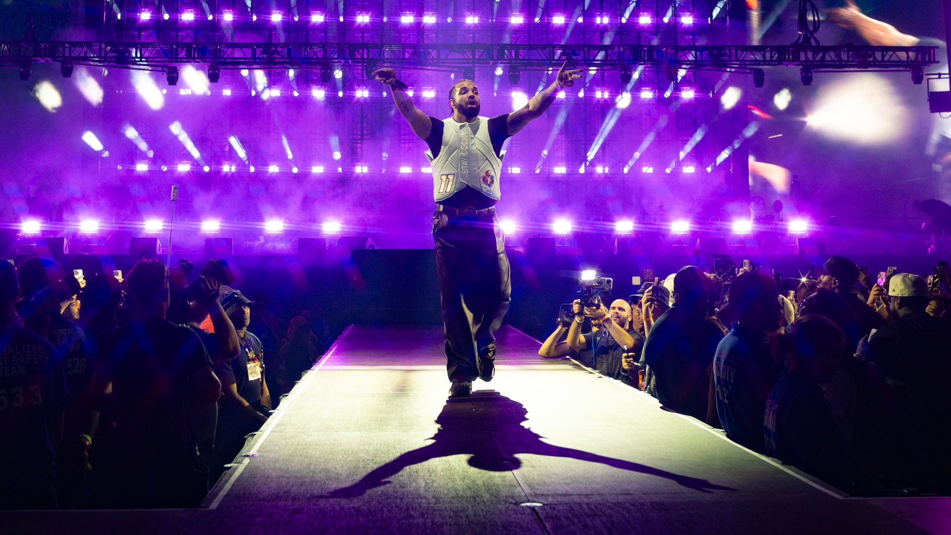 Drake on stage with arms outstretched, surrounded by purple lights and an audience, creating an energetic concert atmosphere.