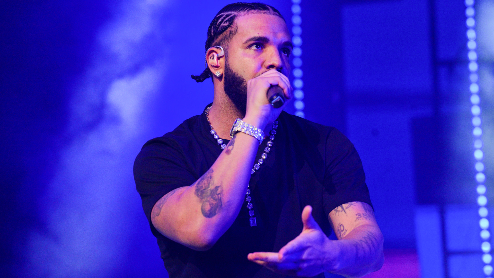 Rapper Drake performing on stage, wearing a black shirt and necklace, holding a microphone with a blue-lit background.