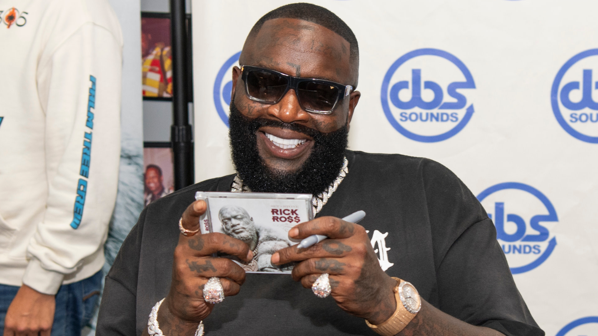 Rick Ross, wearing sunglasses and jewelry, holds a CD with his name and image at a signing event, smiling broadly.