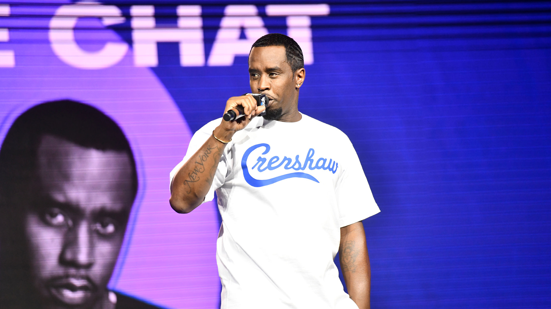 Sean "Diddy" Combs on stage, wearing a white "Crenshaw" shirt, holding a microphone, with a large screen behind him.