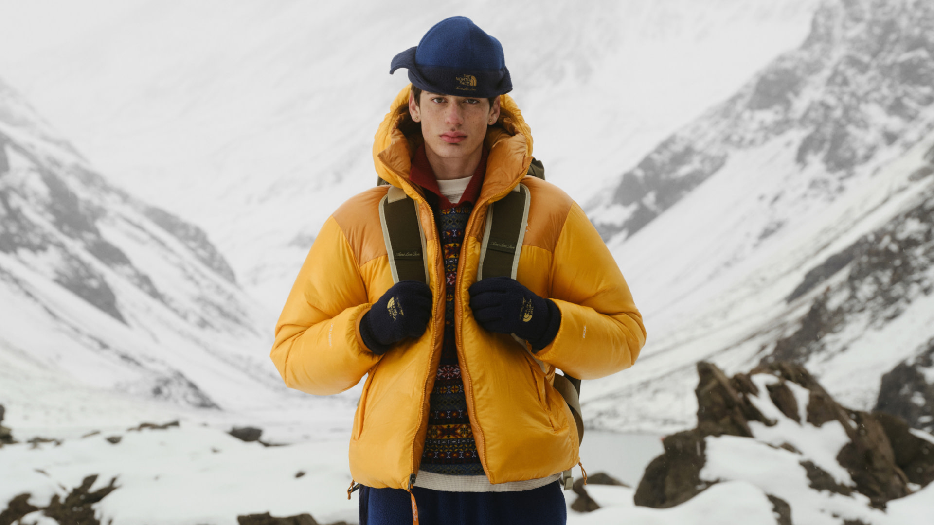 A person in a yellow puffer jacket and blue cap stands in a snowy mountain landscape, wearing gloves and a backpack.