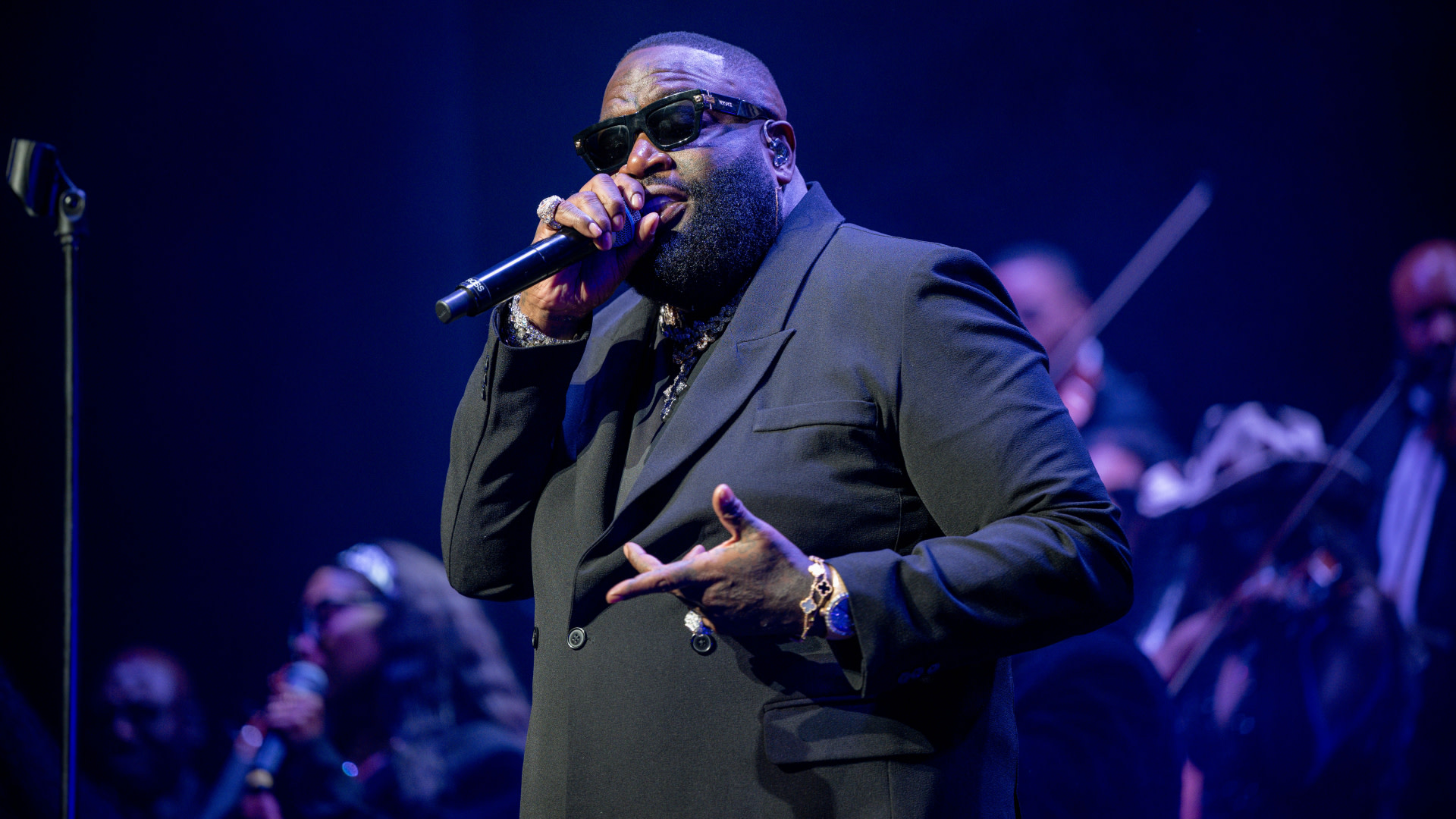 Rapper Rick Ross performing on stage, wearing sunglasses and a black suit, holding a microphone.