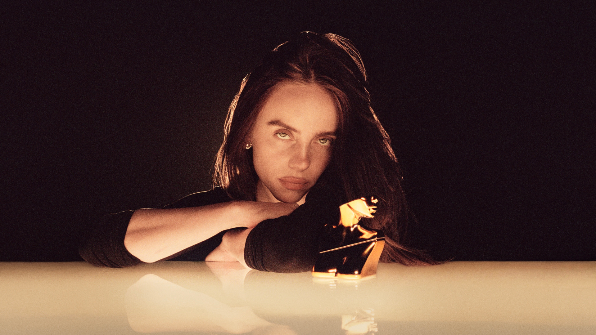 Billie Eilish with long hair leans on a reflective surface, gazing intently. A golden object is in front of her. Dark background.