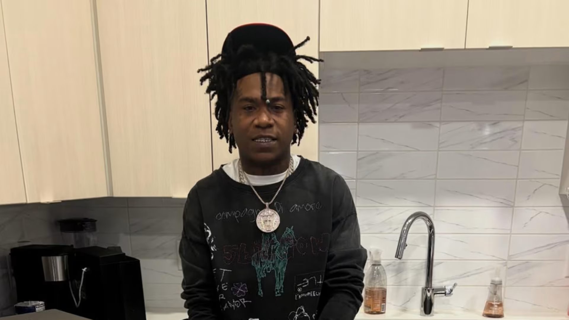 NBA Ben10 with dreadlocks wearing a black graphic sweatshirt and a cap stands in a kitchen with light cabinets and a marble backsplash.