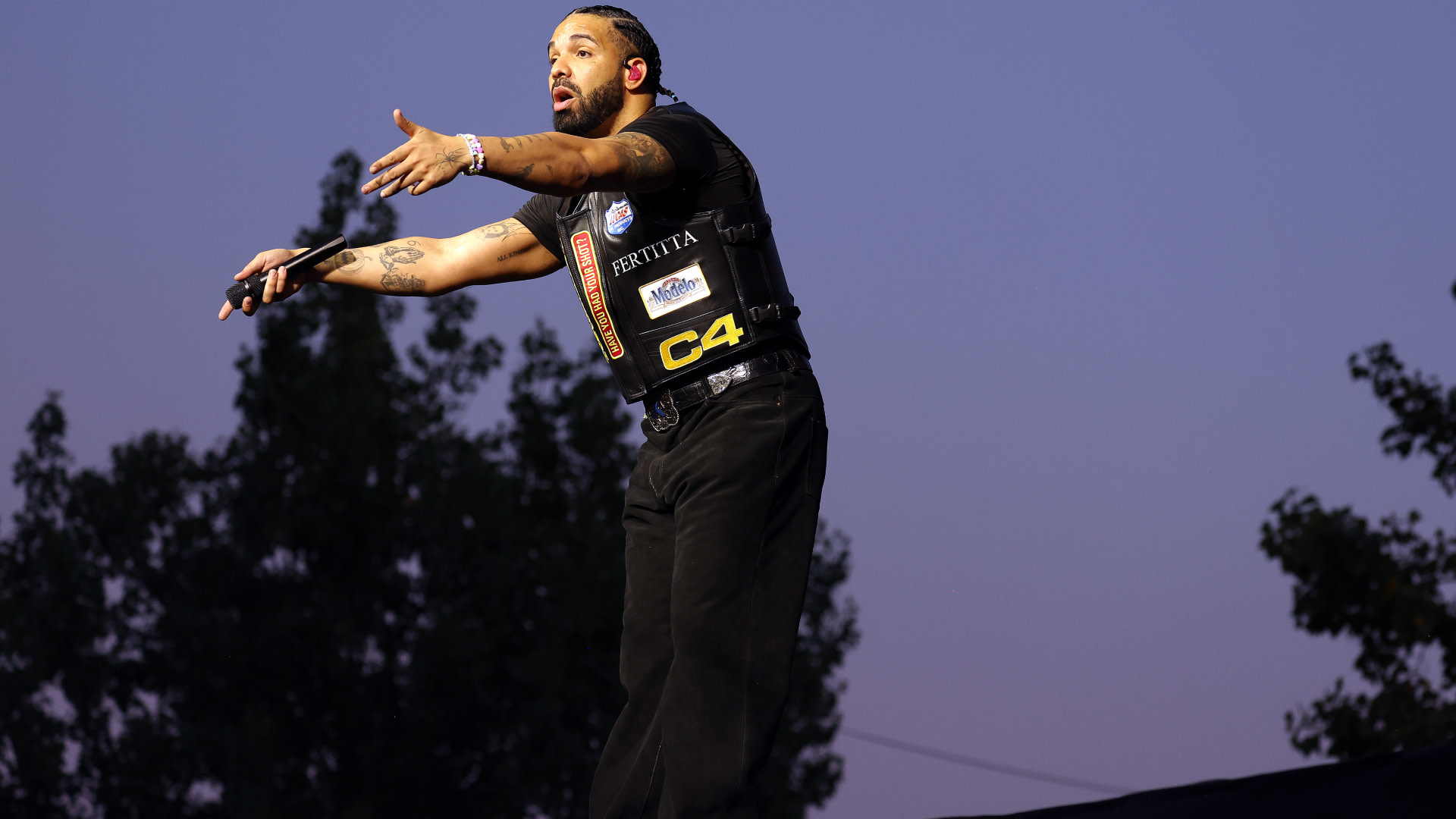 Drake performing on stage, wearing a black outfit with patches, gesturing with his hands, against a twilight sky backdrop.