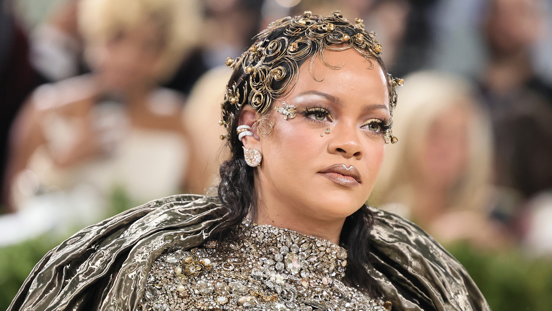 Rihanna at a glamorous event, wearing an ornate headpiece and embellished outfit, with intricate makeup and jewelry.