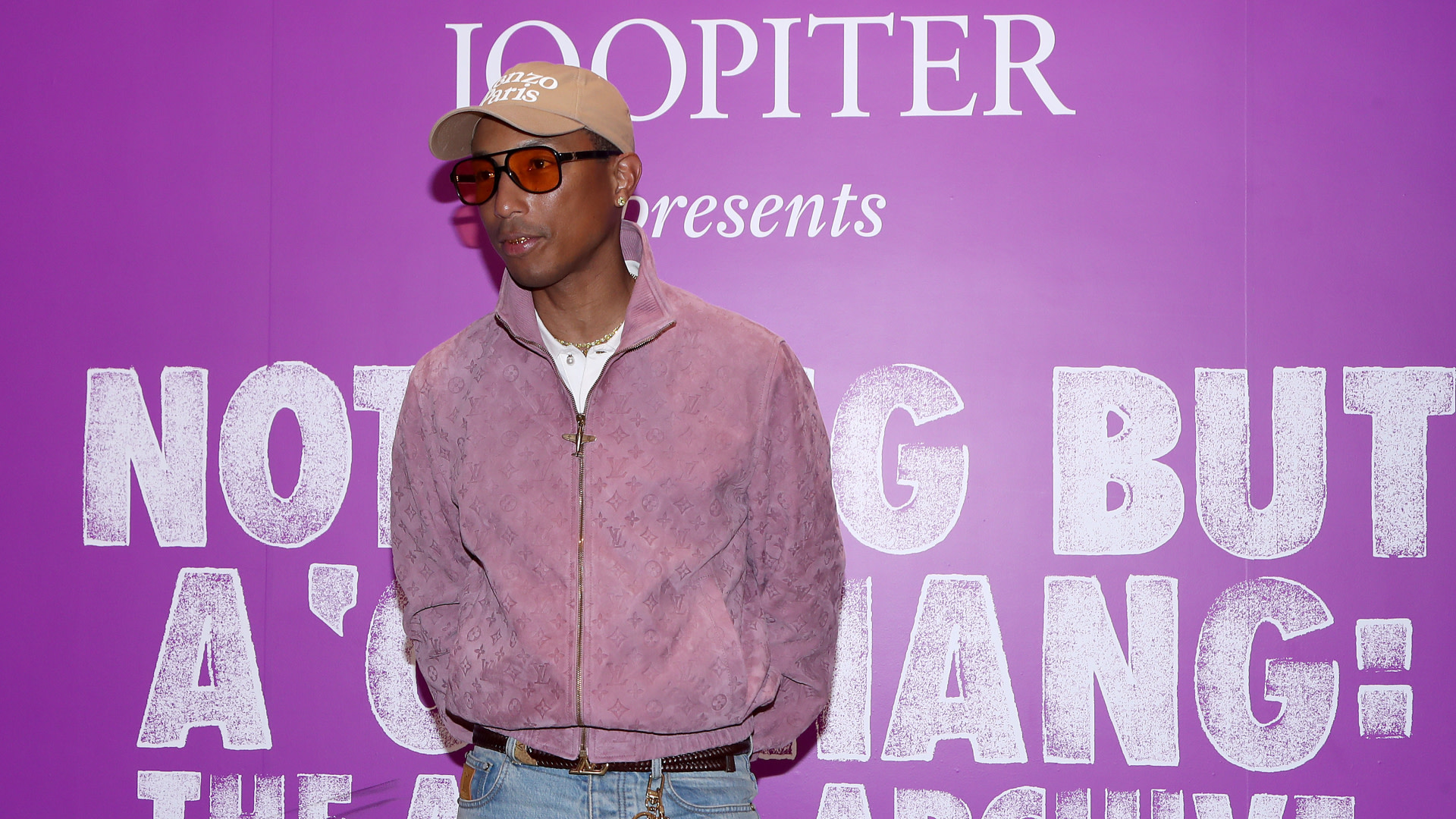 Pharrell Williams in a pink jacket and cap, standing in front of a purple backdrop with white text.
