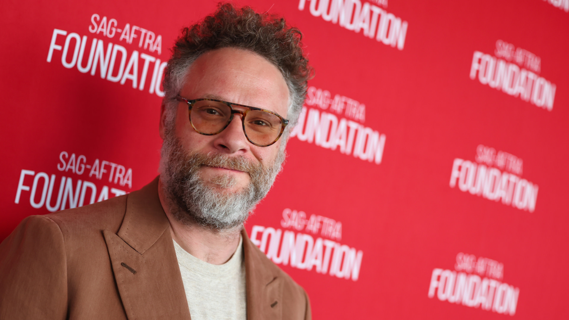 Seth Rogen at a SAG-AFTRA Foundation event, wearing glasses and a brown suit, standing against a red backdrop with the foundation's logo.