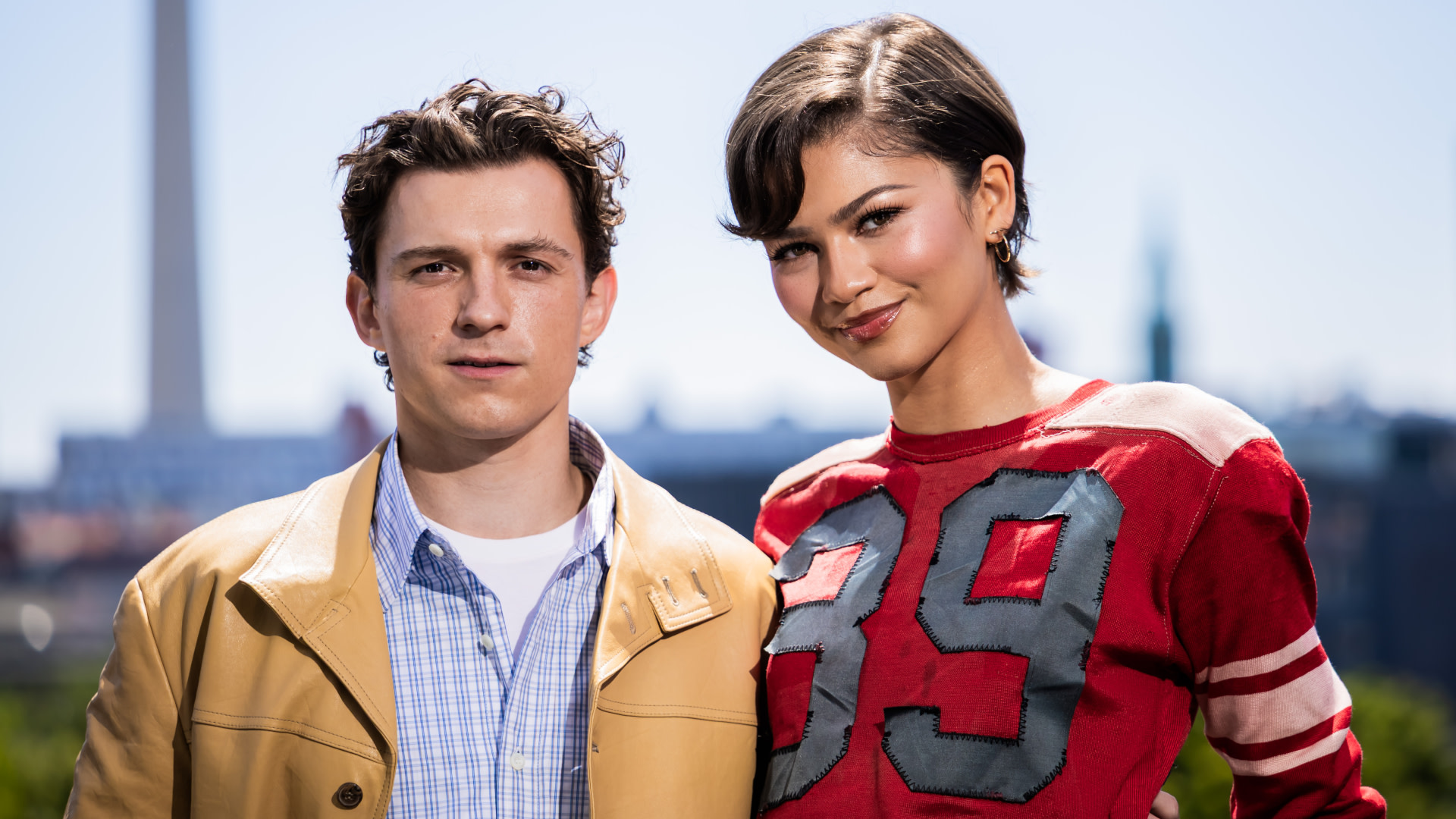 Tom Holland in a tan jacket and Zendaya in a red sweater, standing outdoors with a blurred cityscape background.