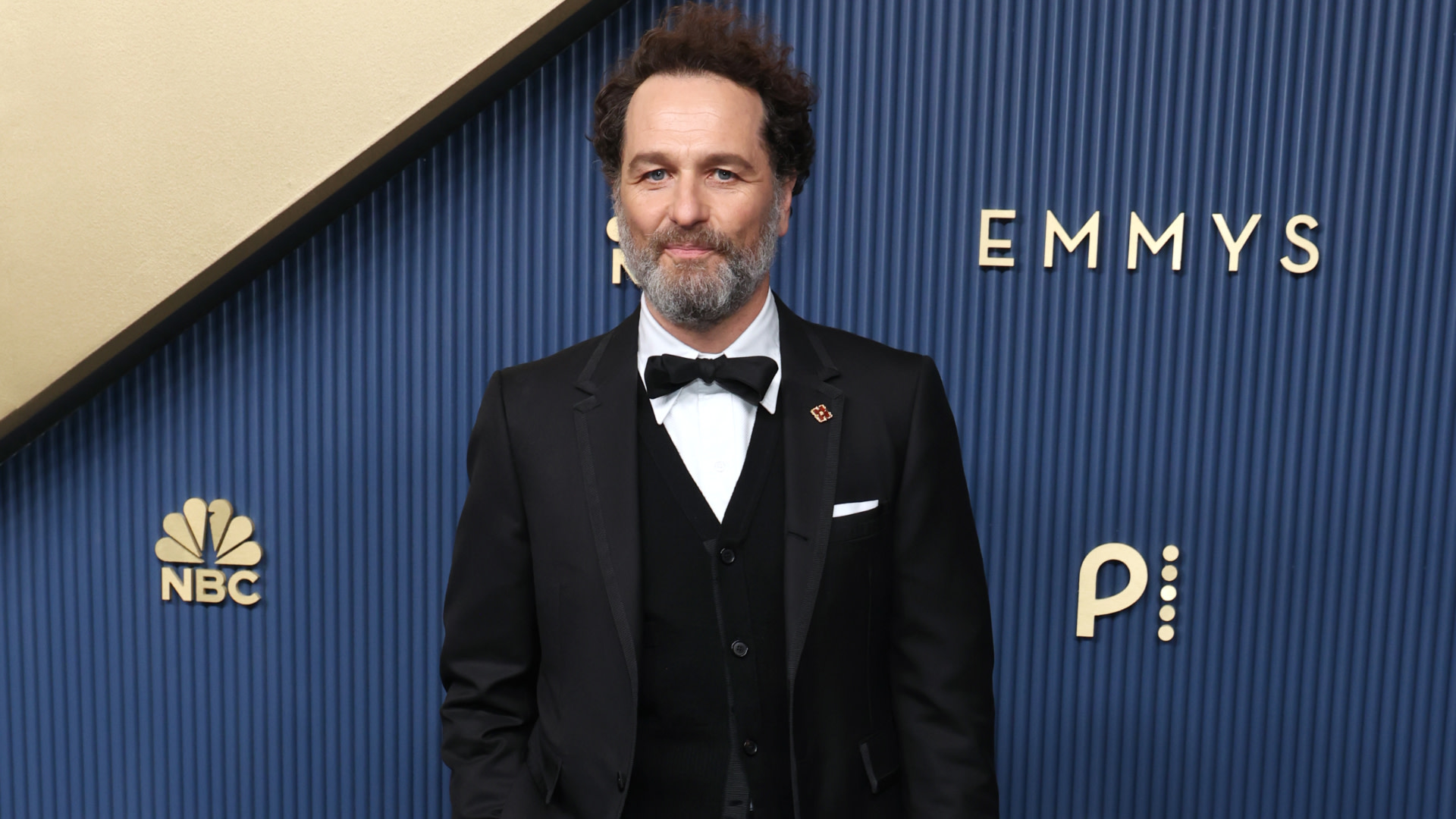 Matthew Rhys in a black tuxedo with a bow tie stands in front of a backdrop with "Emmys" and NBC logos.