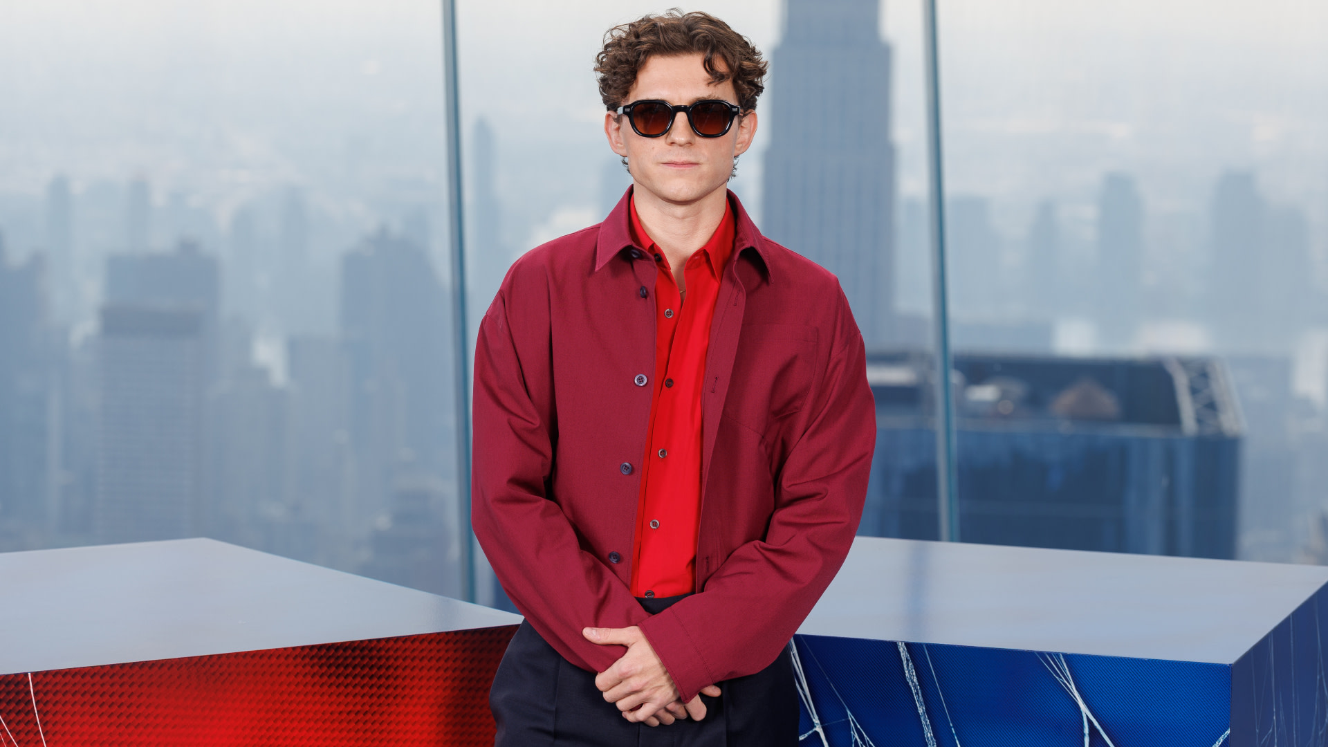Tom Holland in a red shirt and jacket, wearing sunglasses, stands in front of a city skyline.