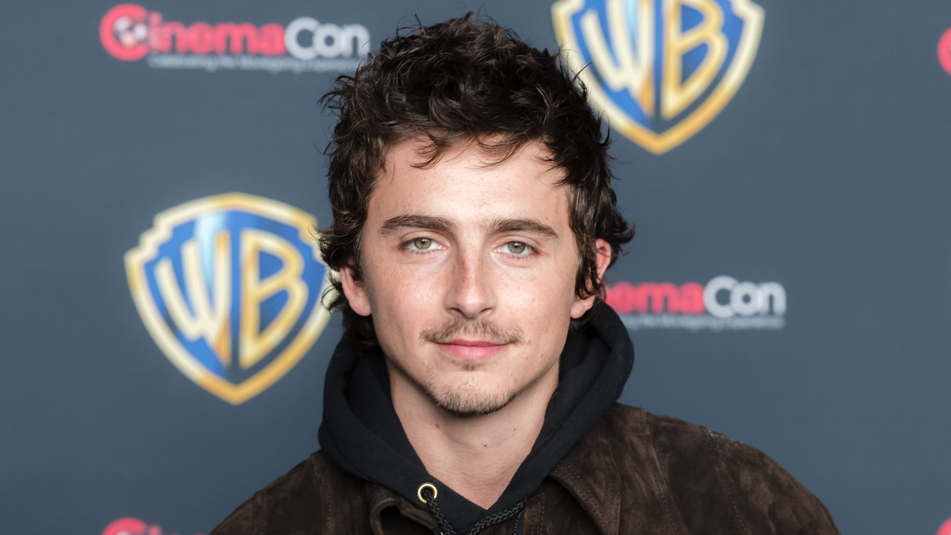 Timothée Chalamet at a Warner Bros. event, wearing a dark jacket and hoodie, with a backdrop featuring the CinemaCon logo.