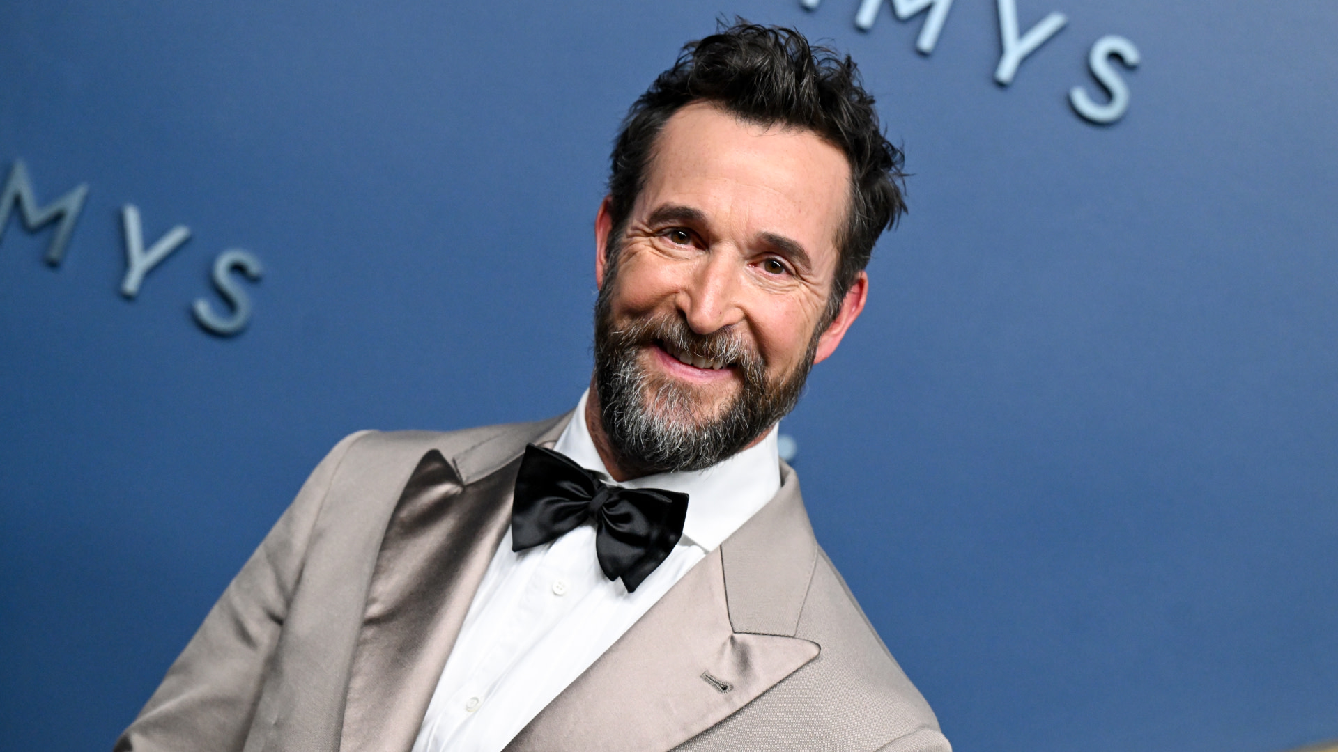 Noah Wyle with a beard and mustache, wearing a light-colored tuxedo and black bow tie, smiles against a blue Emmy Awards backdrop.