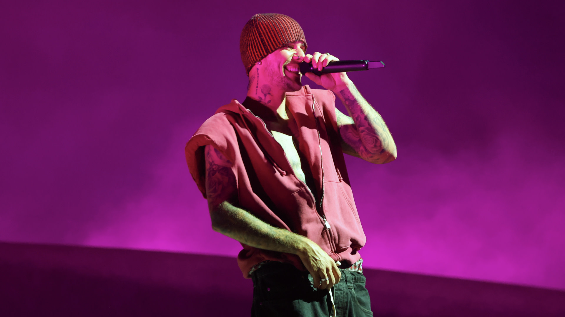 Justin Bieber performs energetically on stage with a microphone, wearing a sleeveless hoodie and beanie, against a vibrant purple background.