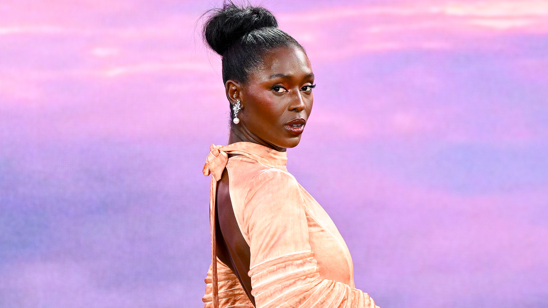 A person in an elegant peach dress poses against a pink and purple gradient background.