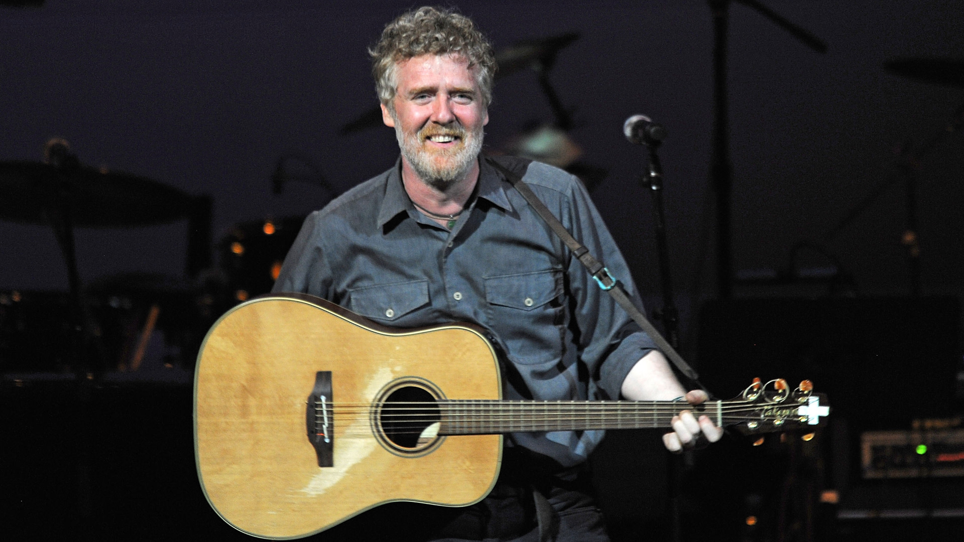 Glen Hansard with a beard, playing an acoustic guitar on stage, smiling, wearing a dark shirt.