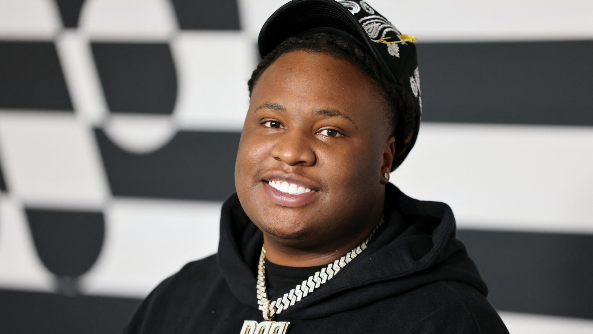 Tay Keith smiling, wearing a black hoodie and cap, with a chain necklace, against a black and white patterned background.