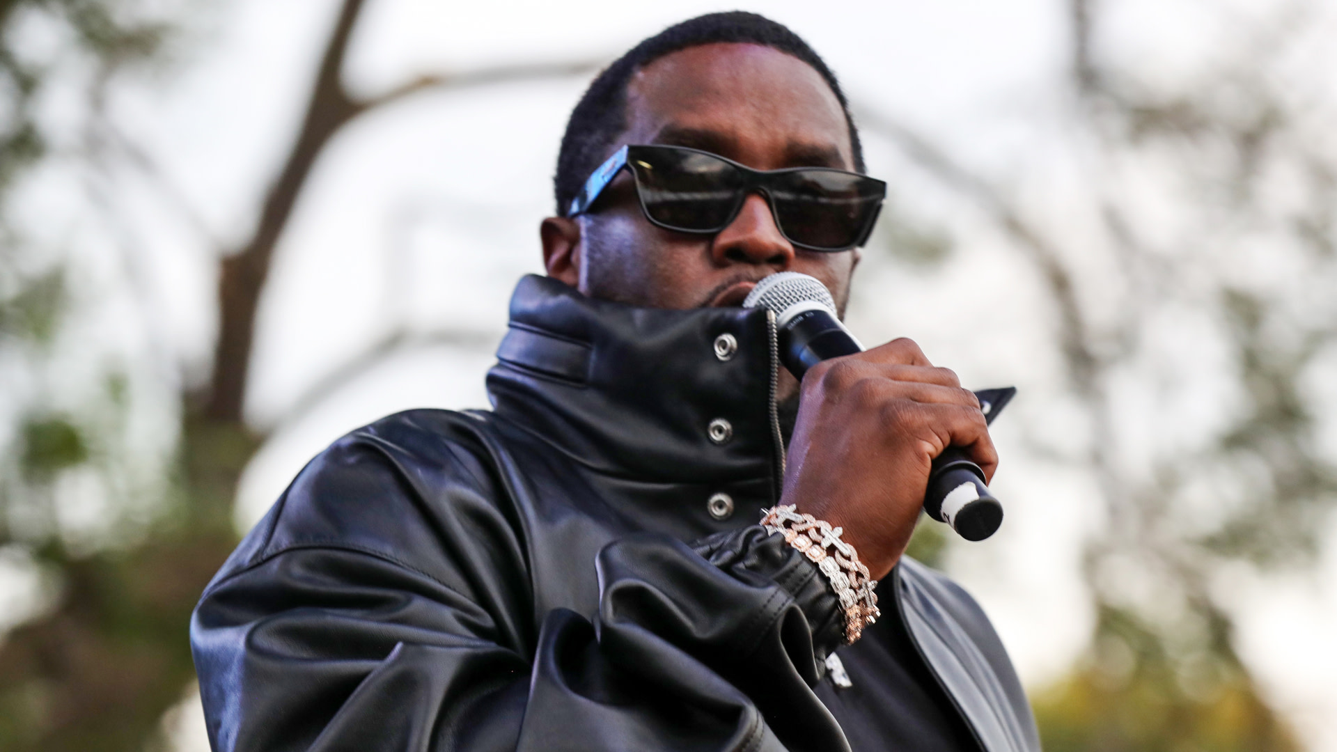 Diddy in a black leather jacket and sunglasses holds a microphone, with trees blurred in the background.