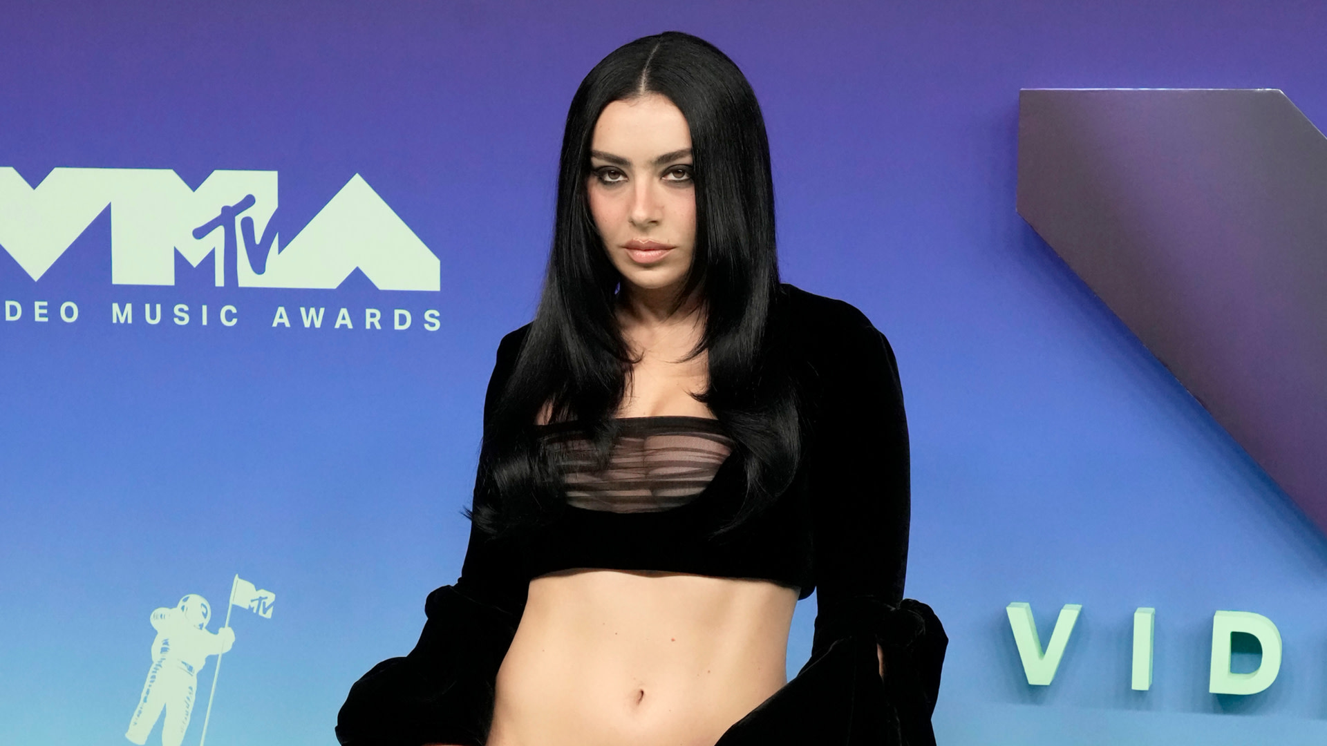 Charli xcx at the MTV Video Music Awards, wearing a black outfit with long sleeves and a sheer top, standing against a blue backdrop.