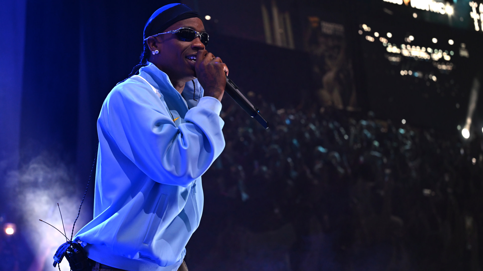 Travis Scott on stage wearing sunglasses and a light blue jacket, holding a microphone with a crowd in the background.