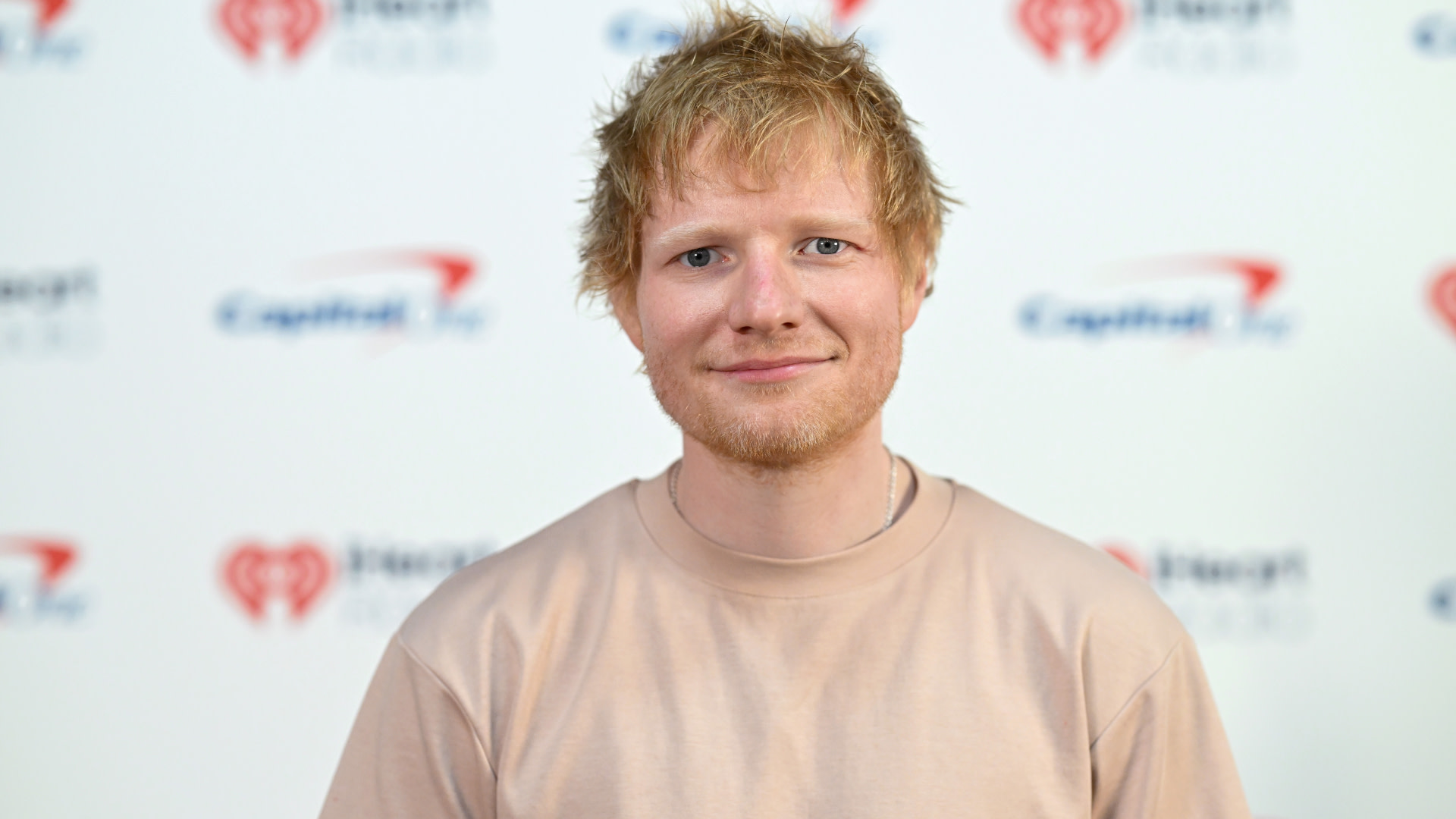 Ed Sheeran smiling, wearing a light pink shirt, with a backdrop featuring iHeartRadio and Capital logos.