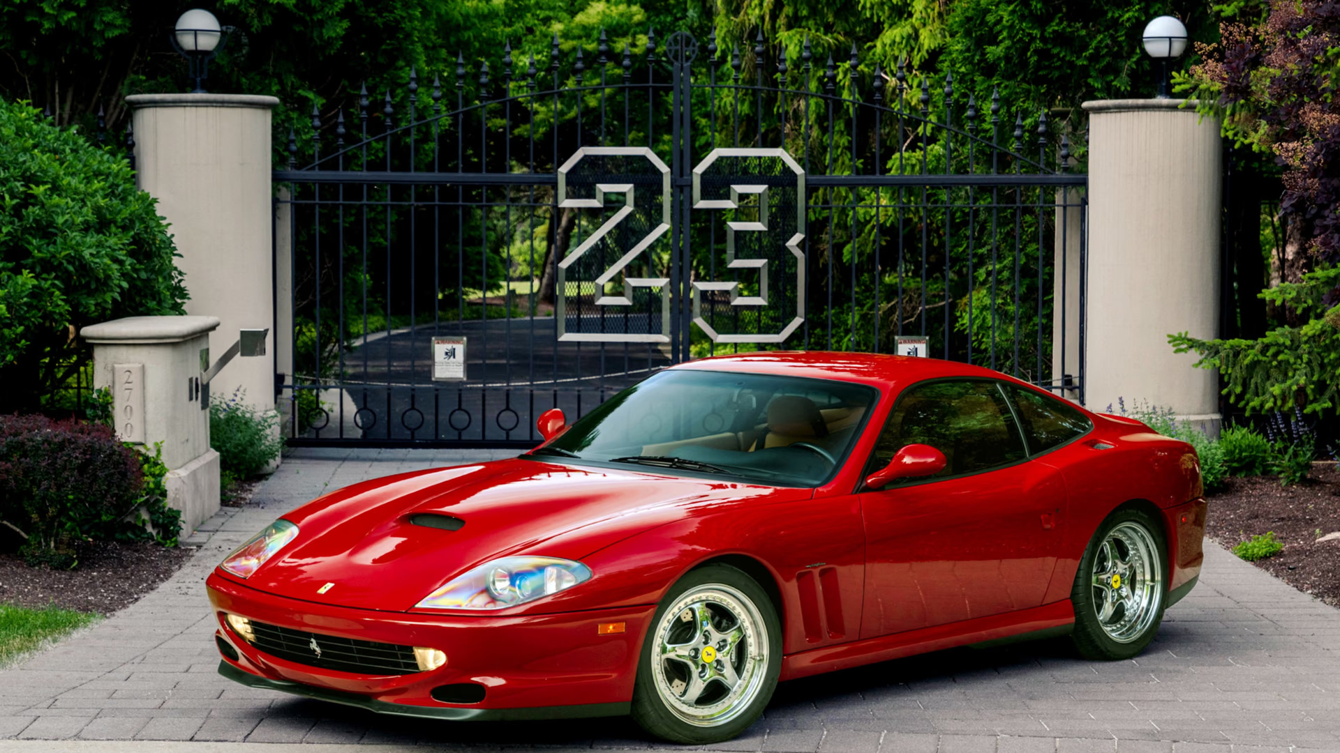 A red Ferrari parked in front of a gated entrance with the number 23. The area is surrounded by lush greenery.