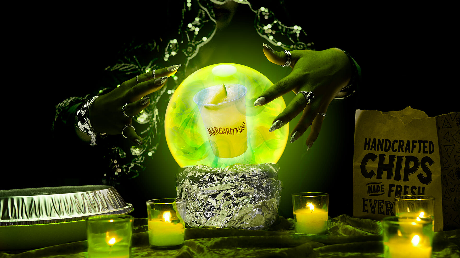 A pair of hands with rings and long nails hover over a glowing crystal ball containing a margarita. Surrounding candles and a chips bag.