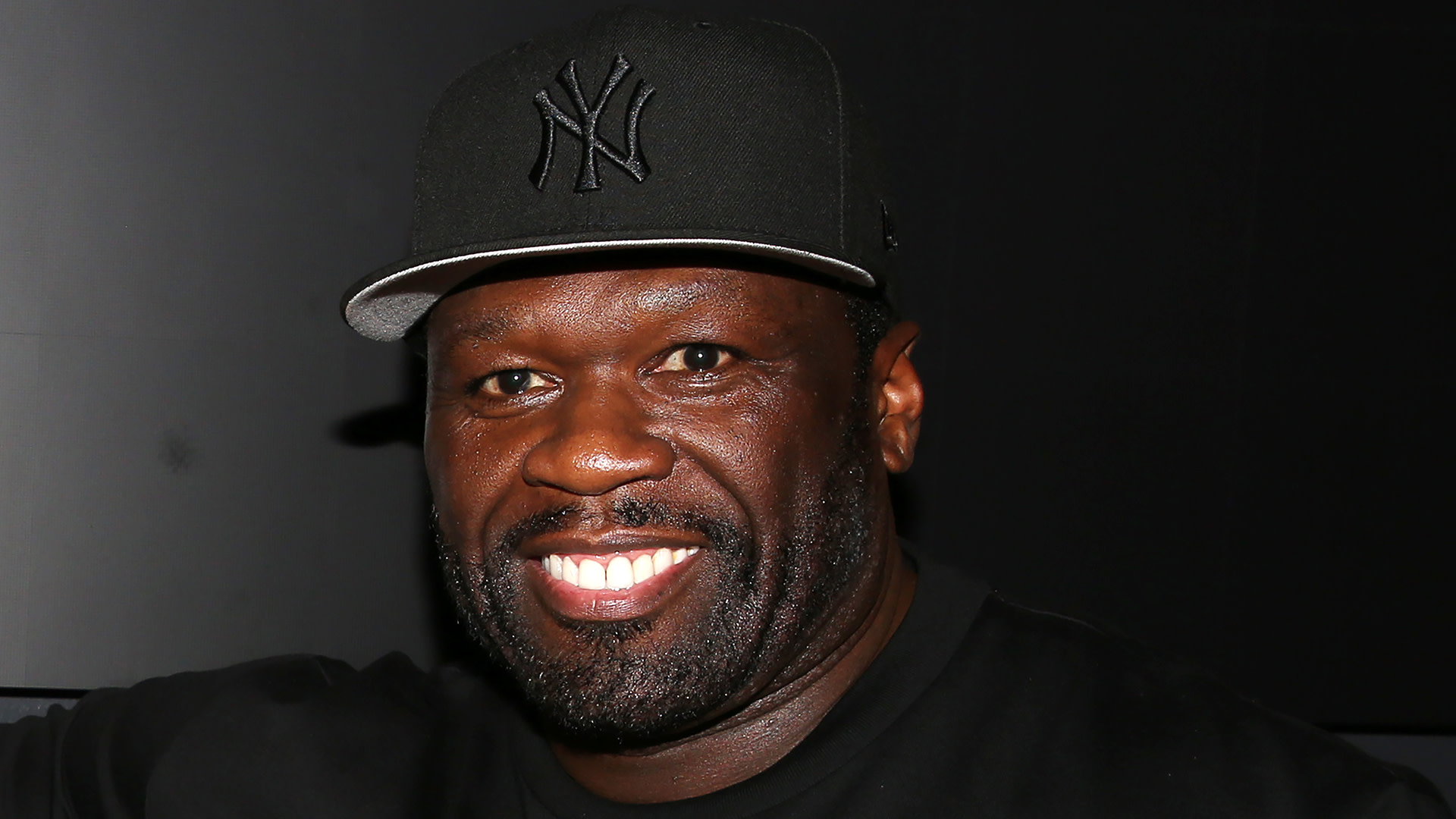 Rapper 50 Cent smiling, wearing a black New York Yankees cap and black shirt, against a dark background.